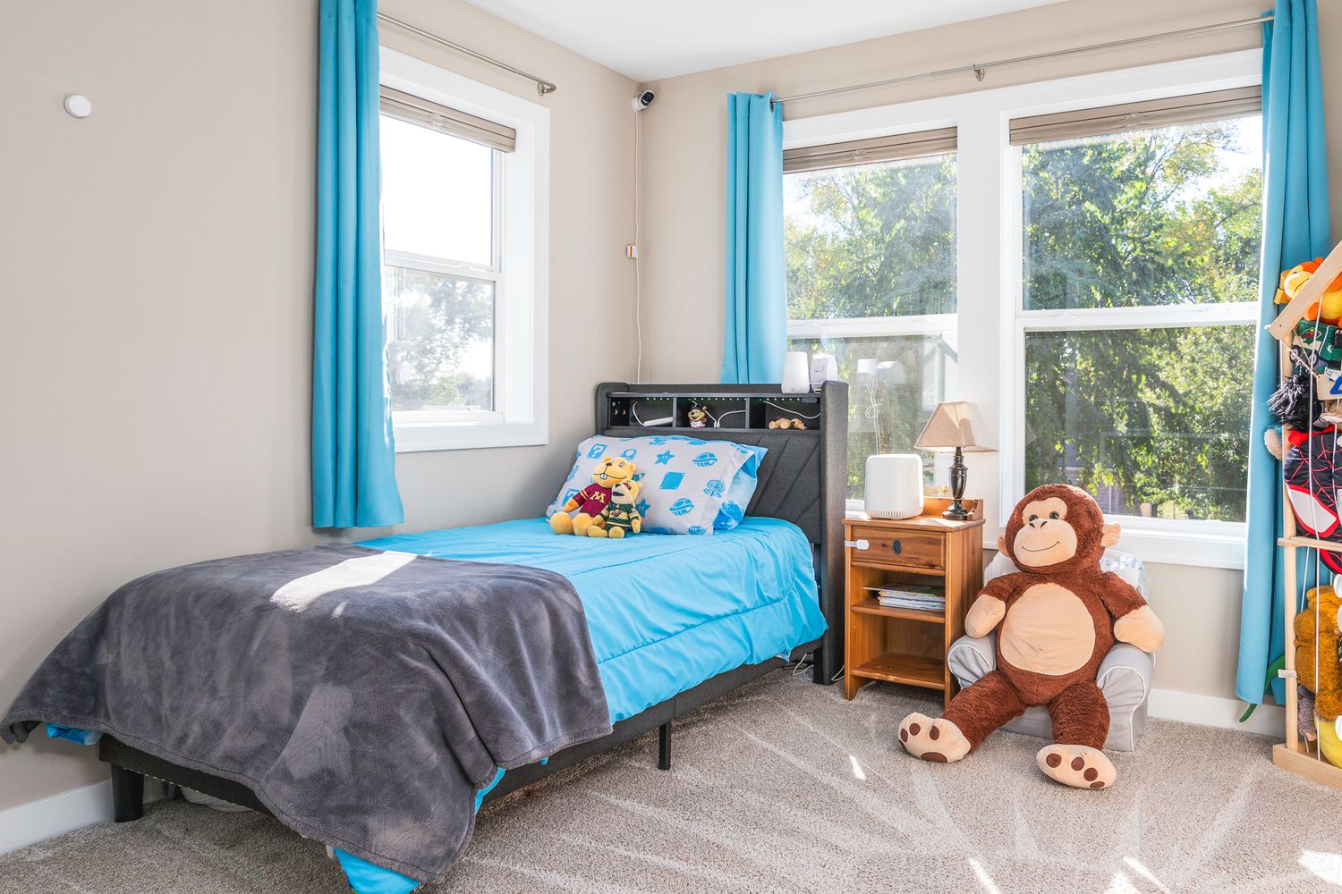 Cheerful kids bedroom with blue curtains, twin bed, stuffed monkey toy, and large windows with natural light.
