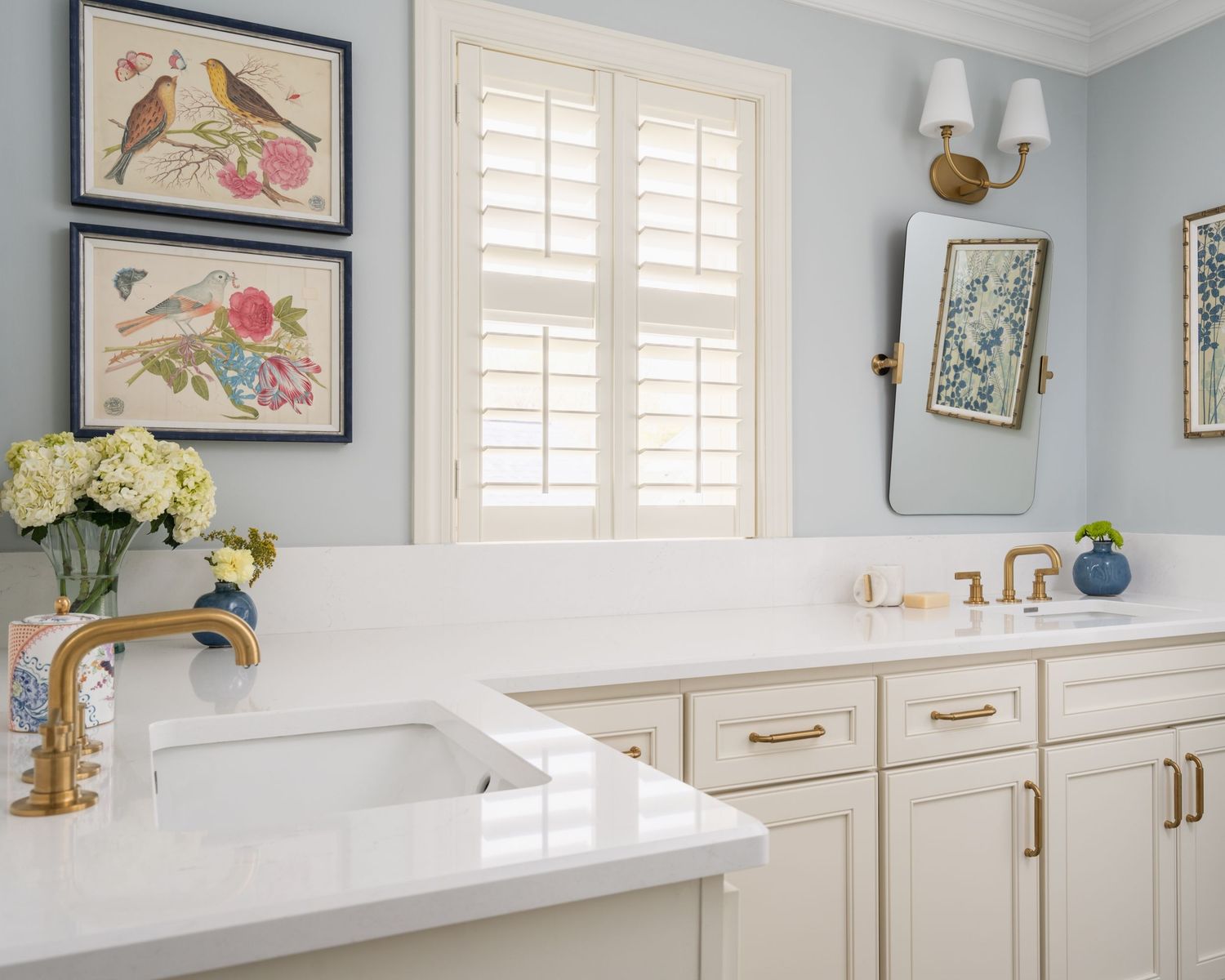 Elegant bathroom with light blue walls, white cabinetry, gold fixtures, botanical artwork, and a marble countertop with a farmhouse sink.