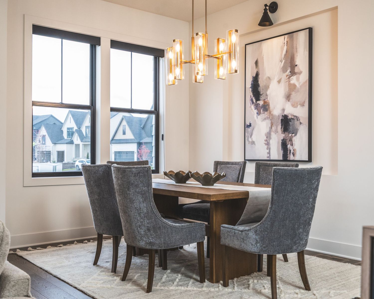 Modern dining room features gray upholstered chairs around a sleek table with pendant lighting and large windows.