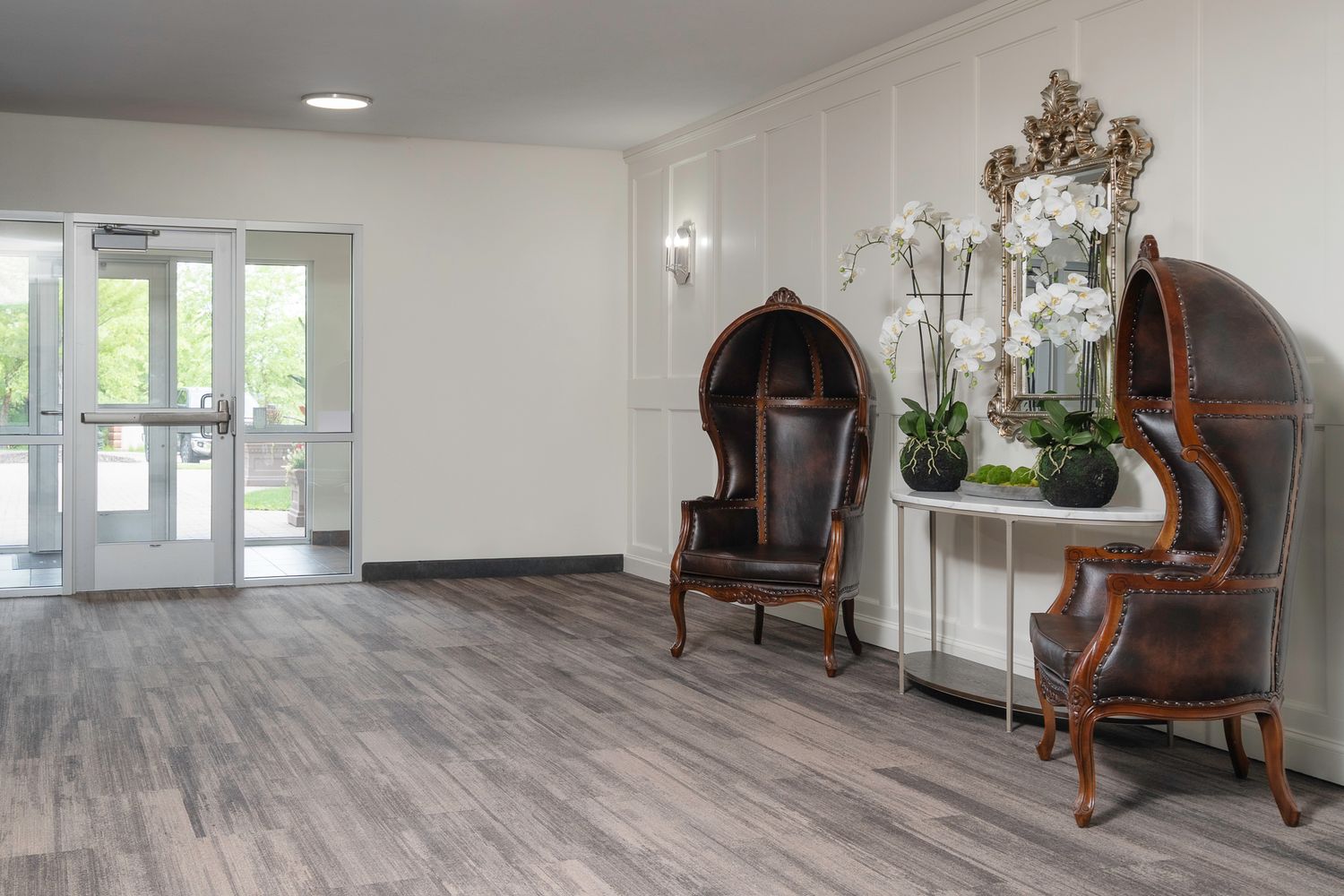 Elegant lobby with ornate canopy chairs, a console table with orchids, and hardwood floors near glass entrance doors.