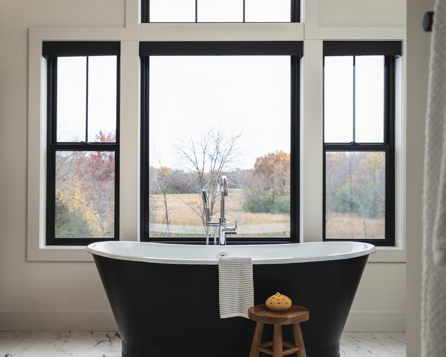 A modern black freestanding bathtub sits beneath large windows overlooking an autumn landscape with a small wooden stool nearby.