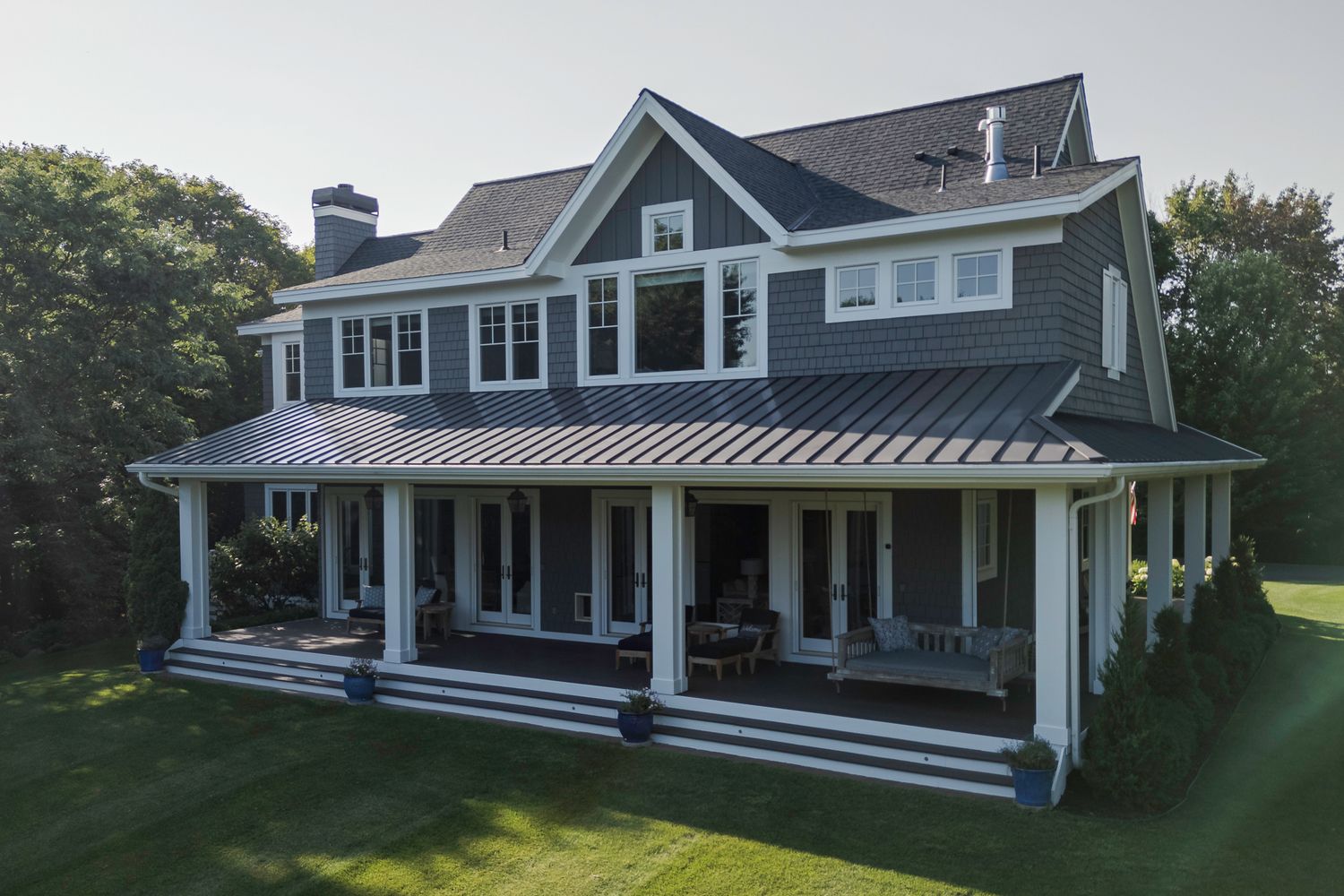 Large two-story craftsman home with dark gray siding, metal roof, wrap-around porch, and lush green lawn.