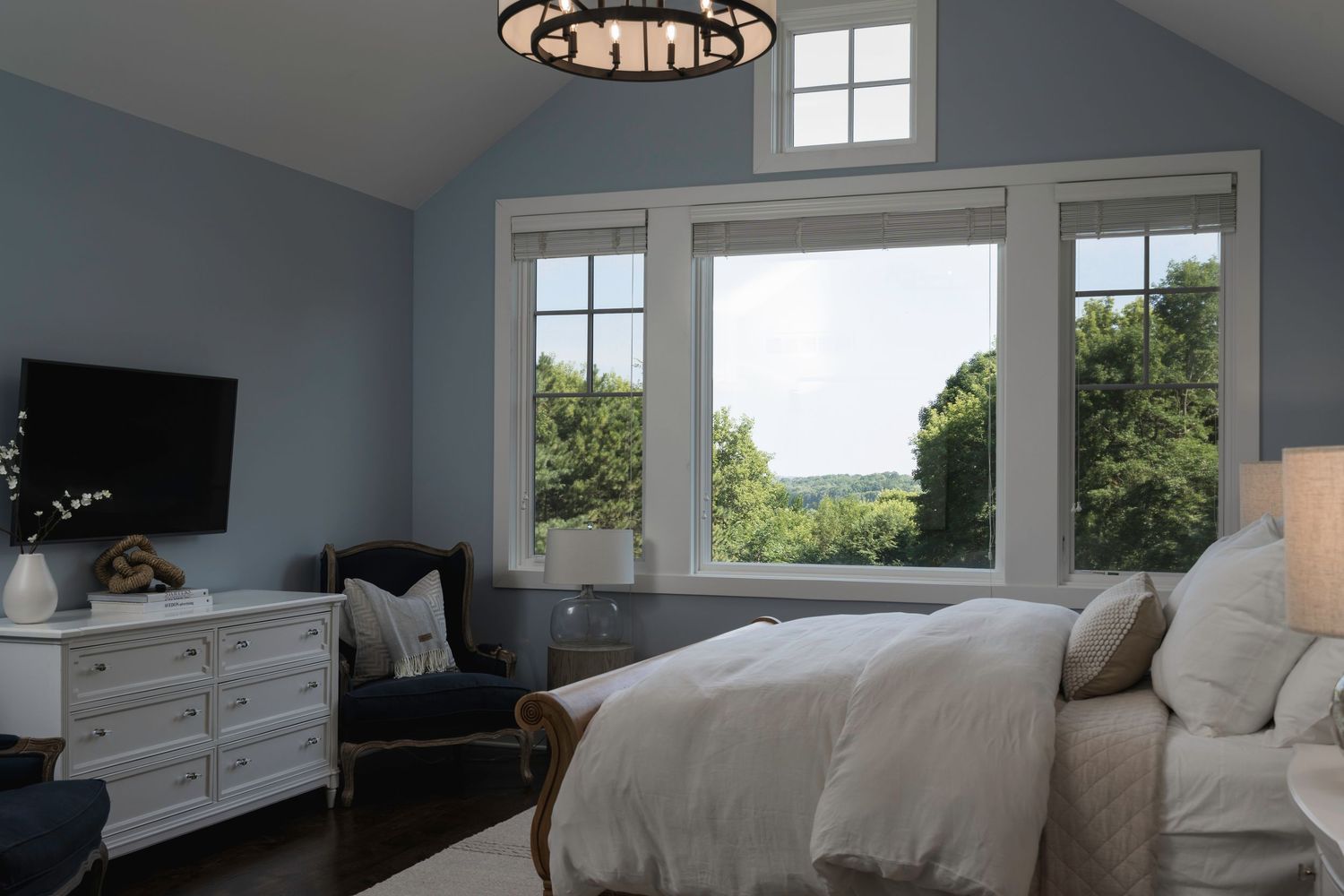 Serene master bedroom with vaulted ceiling, large windows, scenic views, white dresser, and neutral bedding.