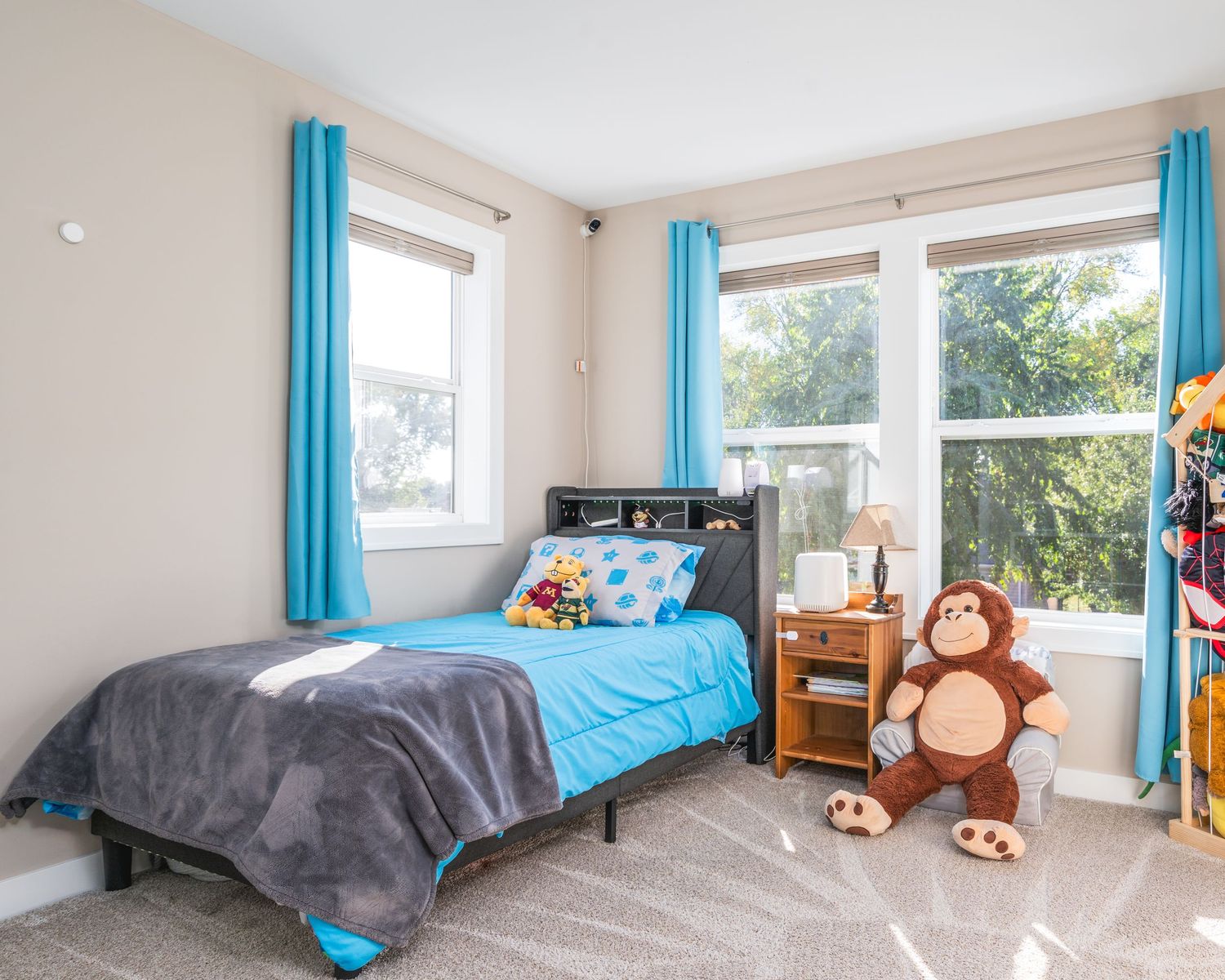 Bright bedroom with turquoise curtains, plush monkey toys, and large windows offering natural light.