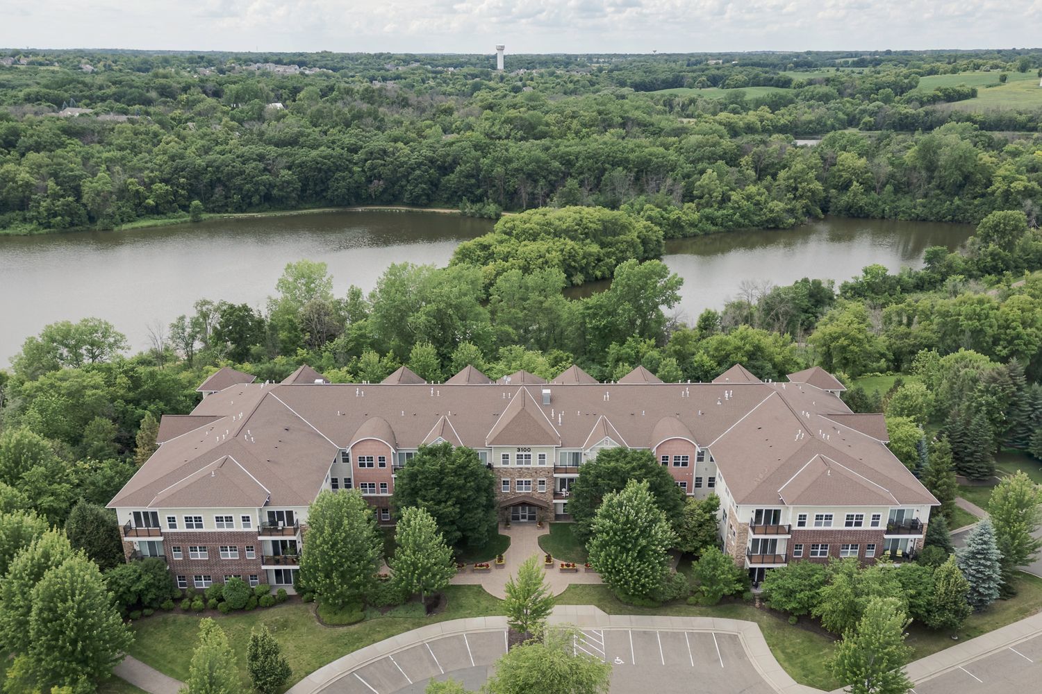 Aerial view of luxury brick apartment complex surrounded by lush trees beside a serene lake in a suburban setting.