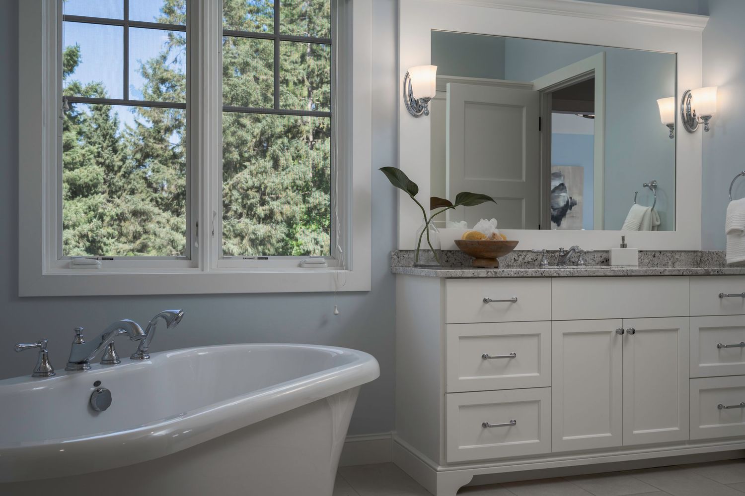 Elegant bathroom with freestanding soaking tub, white vanity with granite countertop, large mirror, and bright window view.