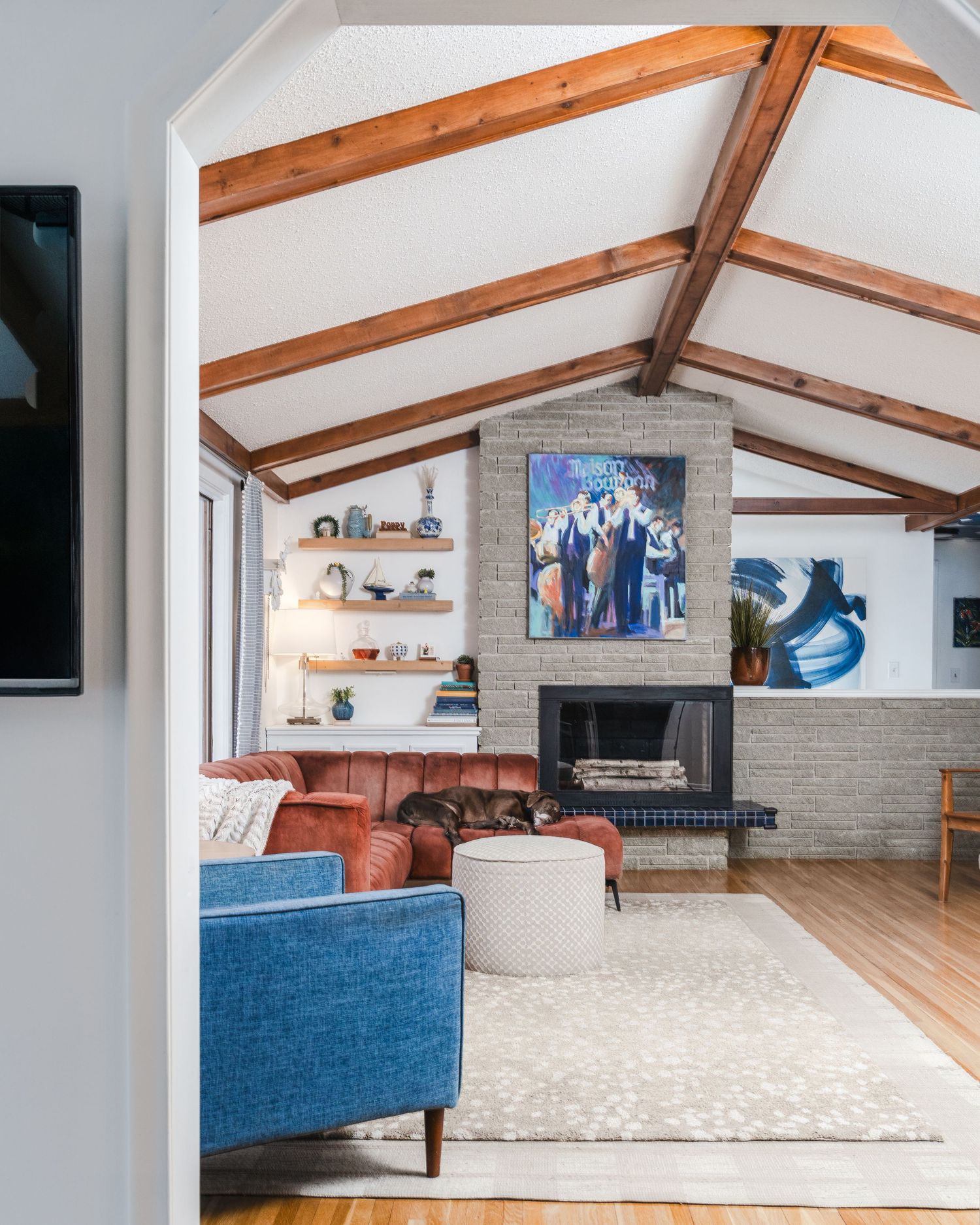 Modern living room with exposed wood beams, blue sofa, leather chairs and floating shelves around a brick fireplace.