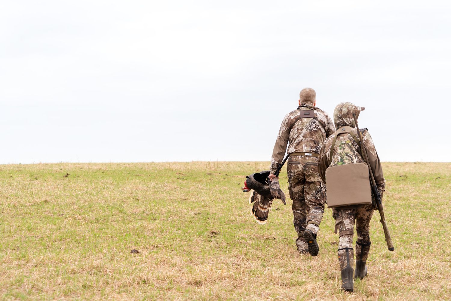 Two hunters in camo walking across an open field carrying hunting gear.