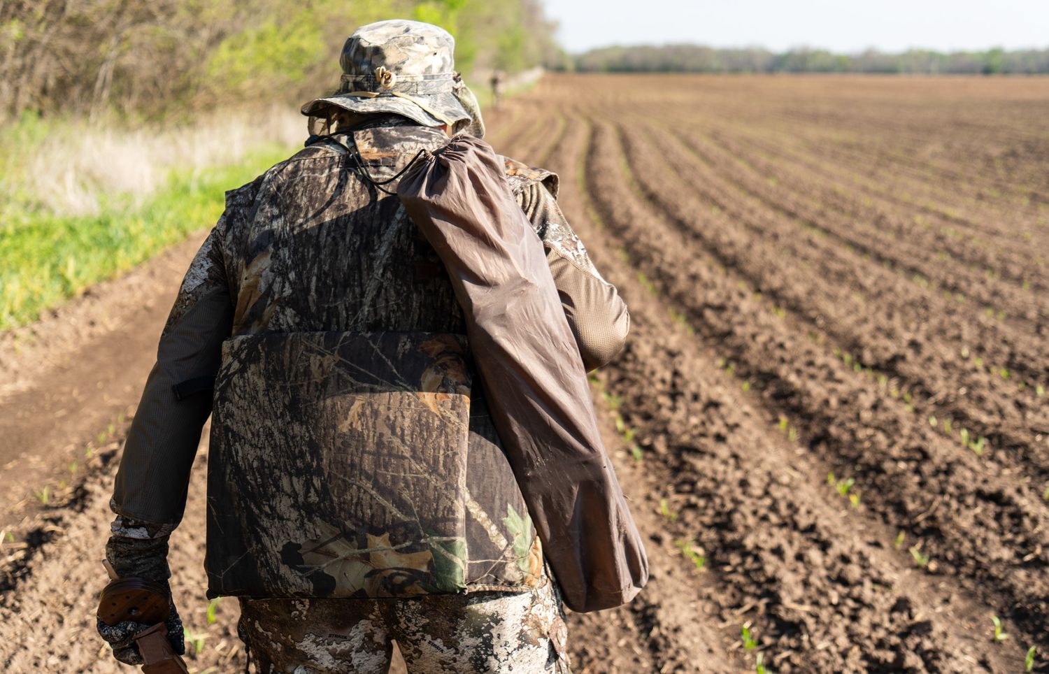 Hunter in camouflage gear walking along the edge of a plowed field.