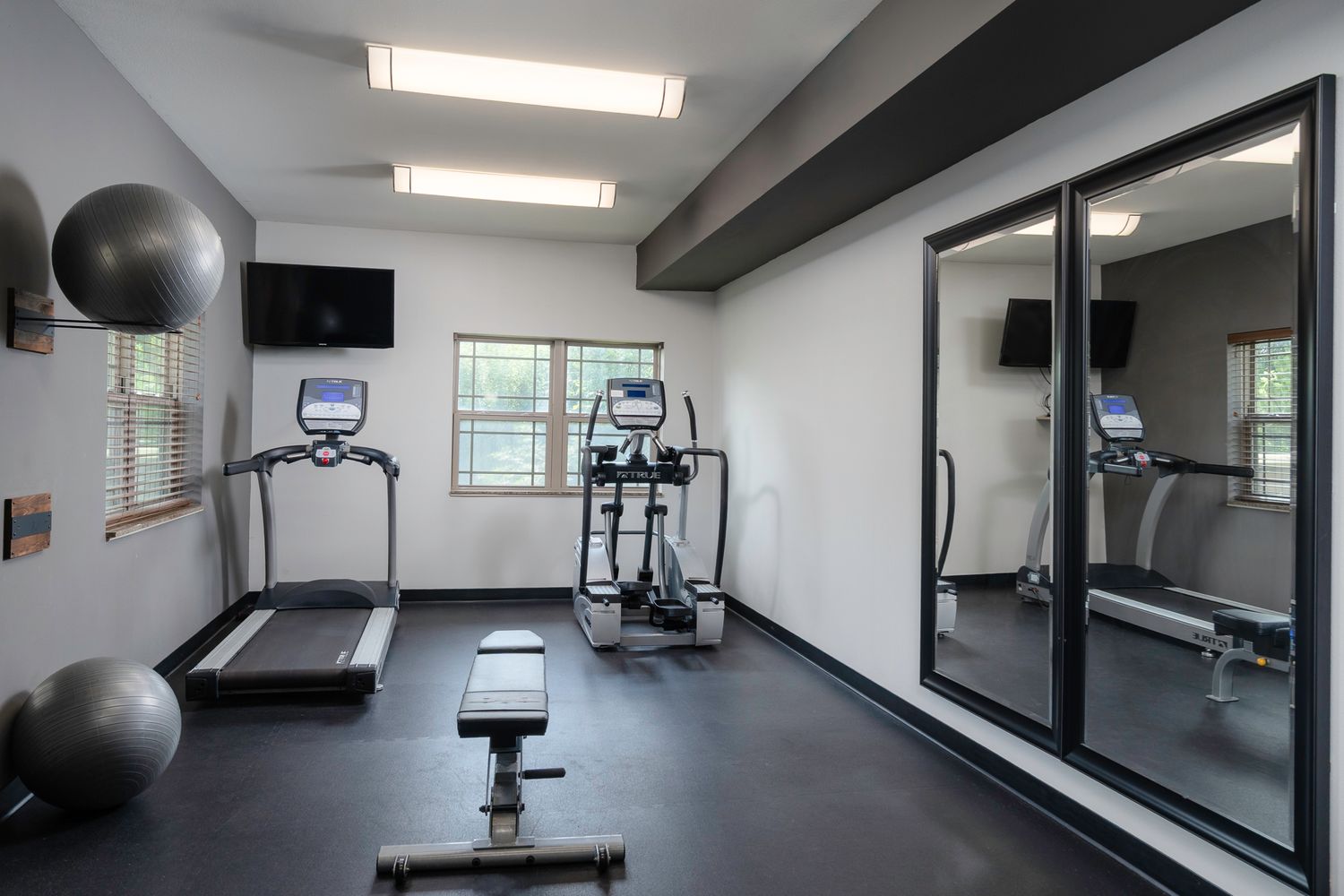 Modern apartment fitness center with treadmill, elliptical, weight bench, exercise balls, and mirrored wall on dark flooring.