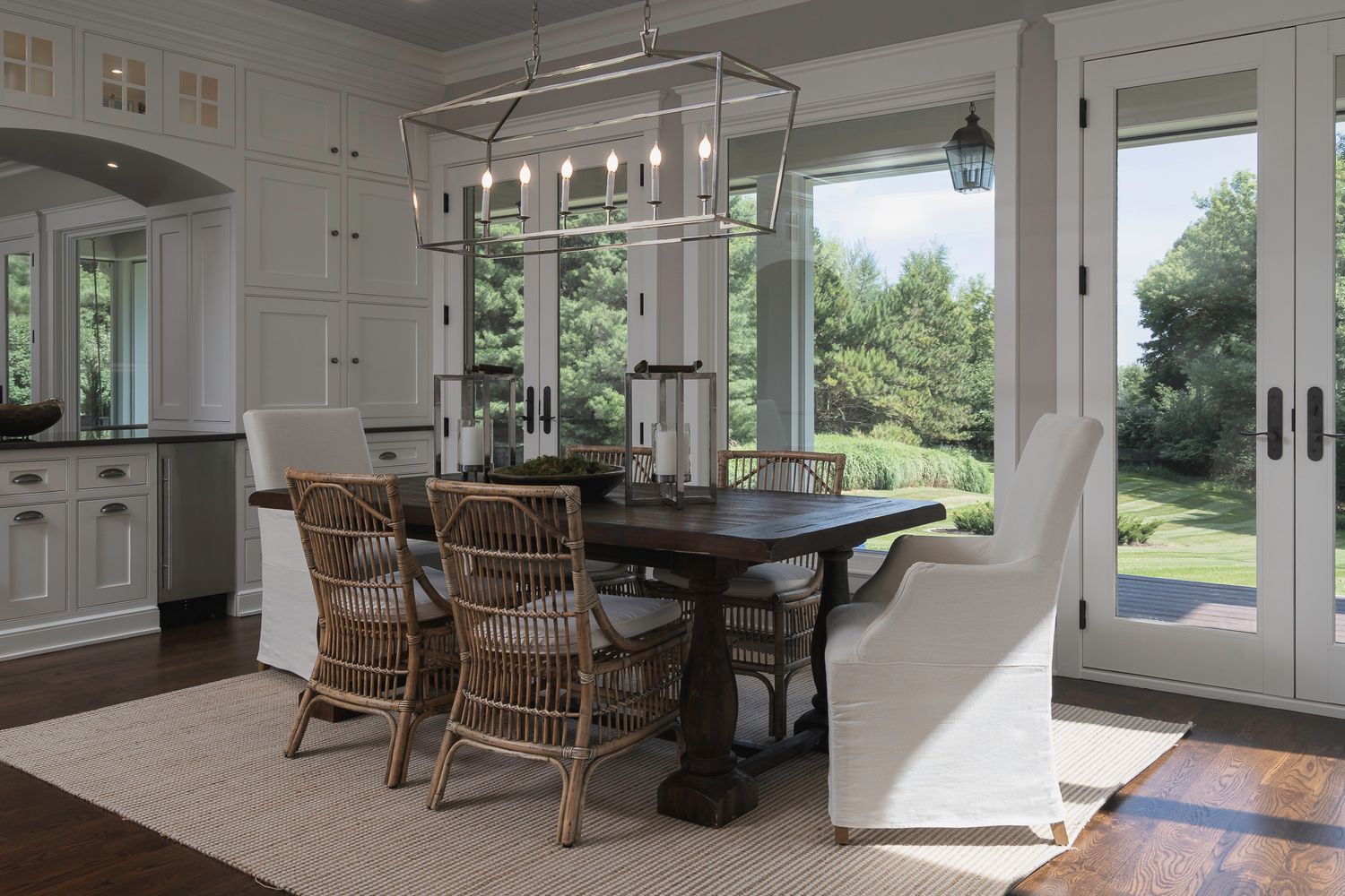 Elegant dining room with dark wood table, wicker chairs, white cabinetry, hardwood floors, and large windows overlooking lush greenery.