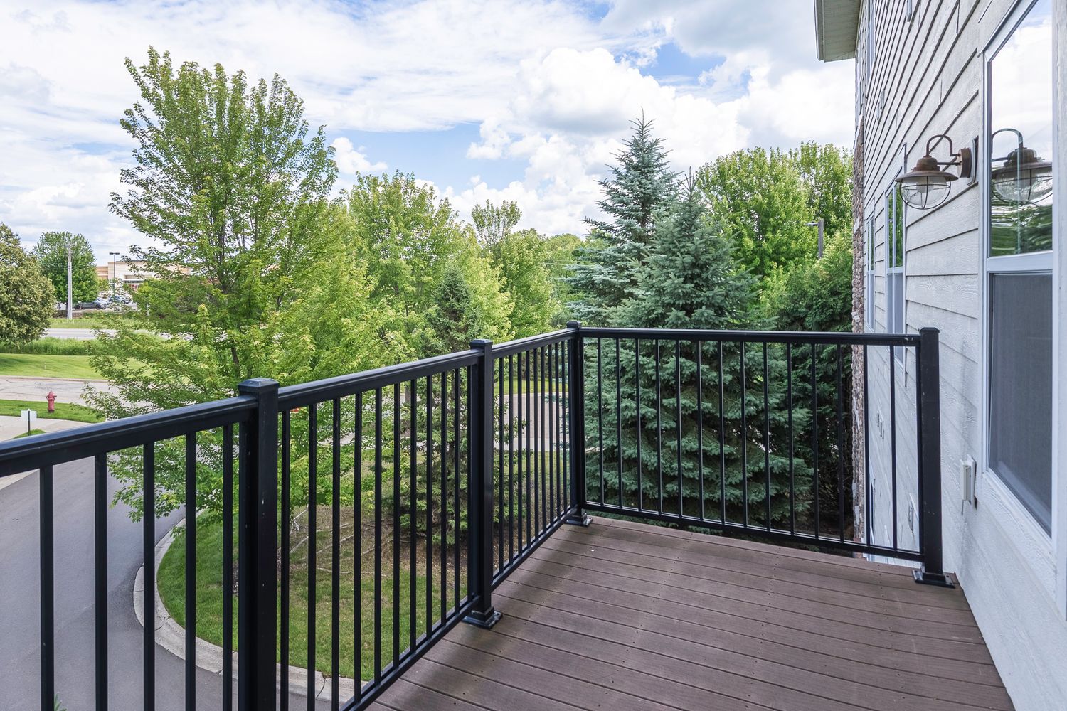 A wooden deck with black metal railings overlooking lush green trees and a suburban neighborhood on a partly cloudy day.