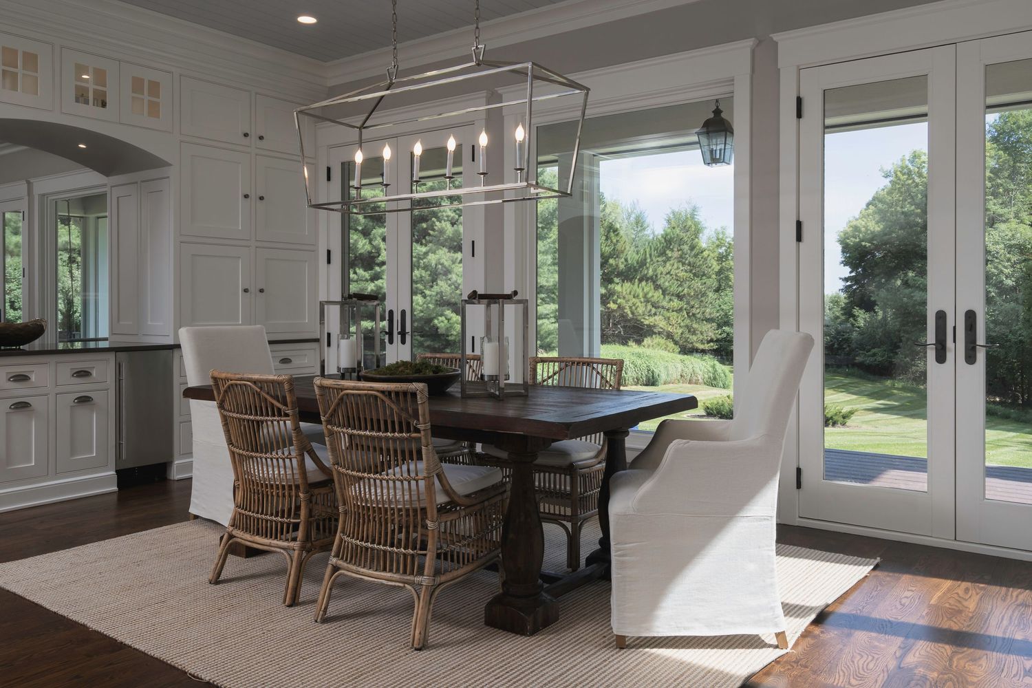 Elegant dining room with wicker chairs, dark wood table, modern chandelier, white built-ins, and French doors overlooking lush greenery.
