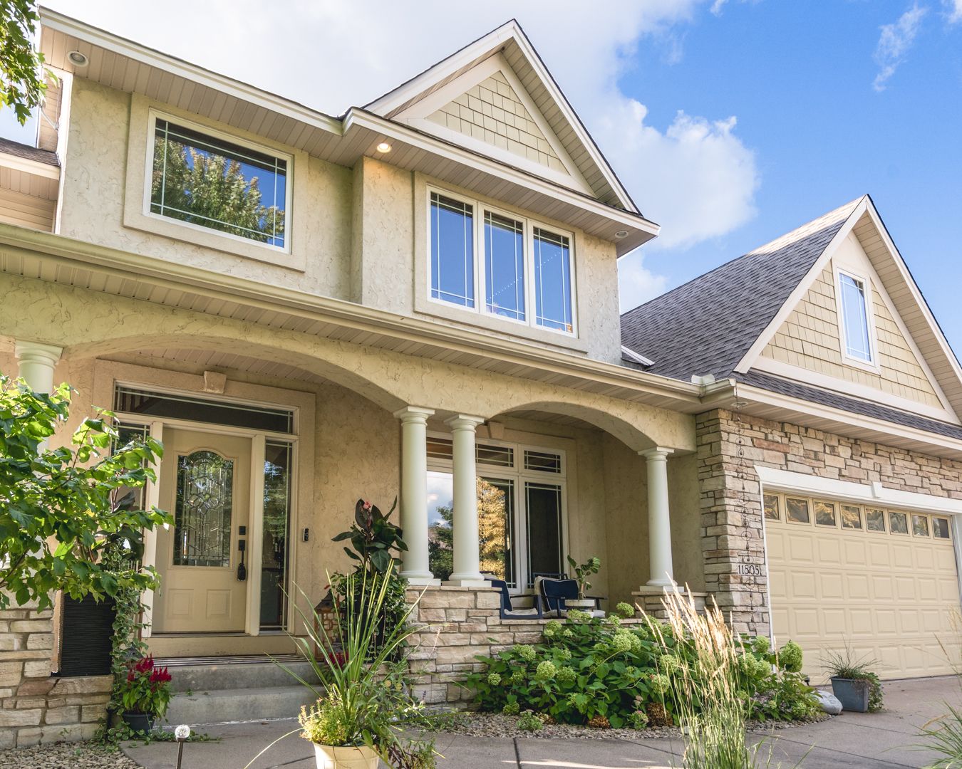 Elegant two-story suburban house with beige siding, stone accents, and a large attached garage under blue summer sky.