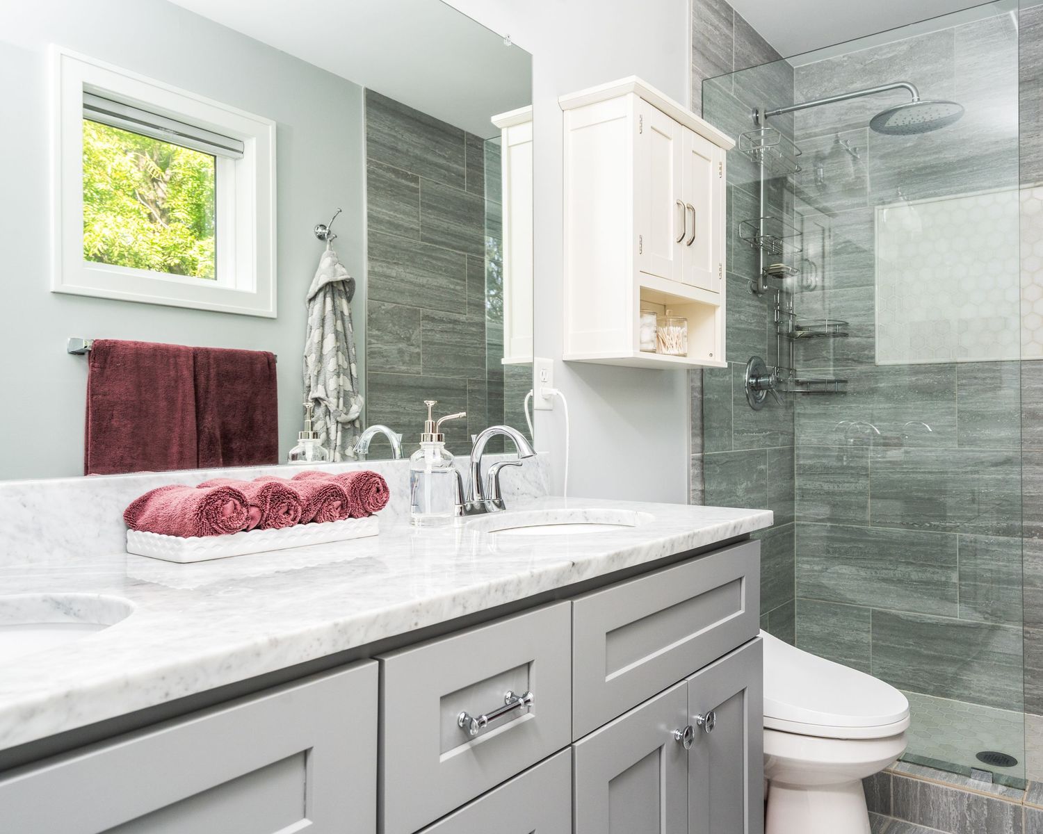 Modern bathroom with gray vanity, marble countertop, glass shower enclosure and burgundy towels.