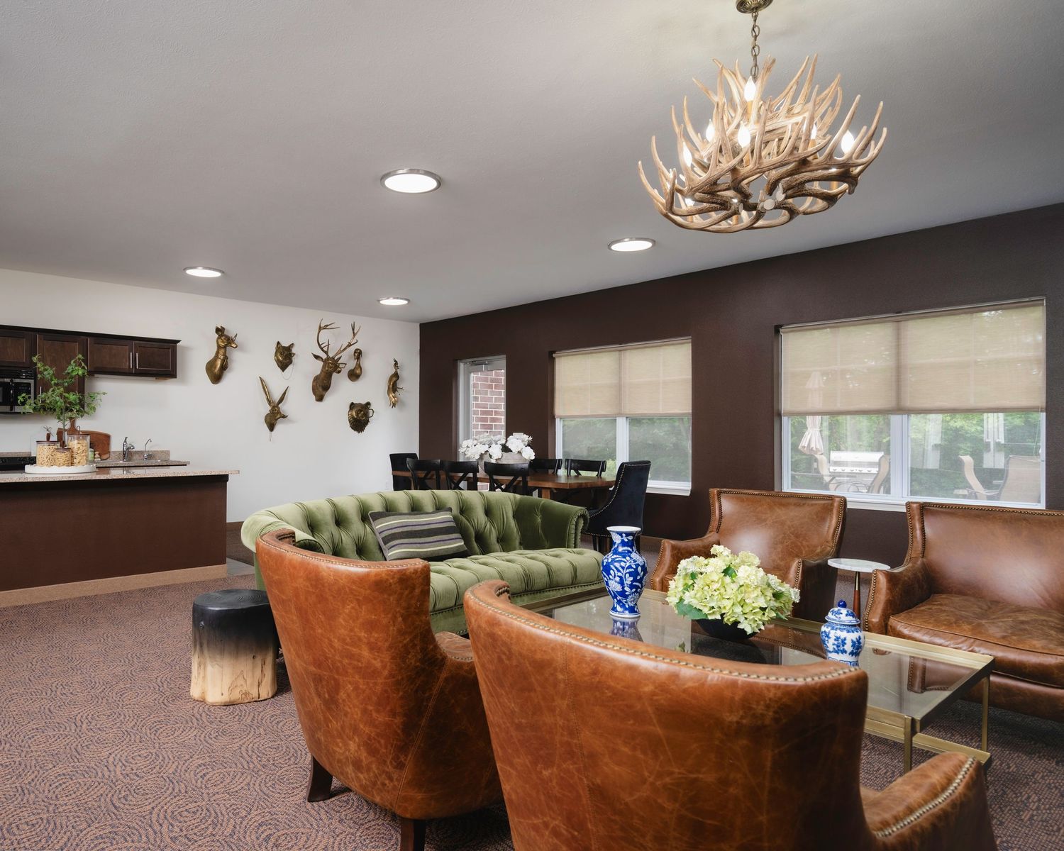 Rustic lodge lounge with leather chairs, green velvet sofa, antler chandelier, animal mounts, and a bar area on brown carpet.