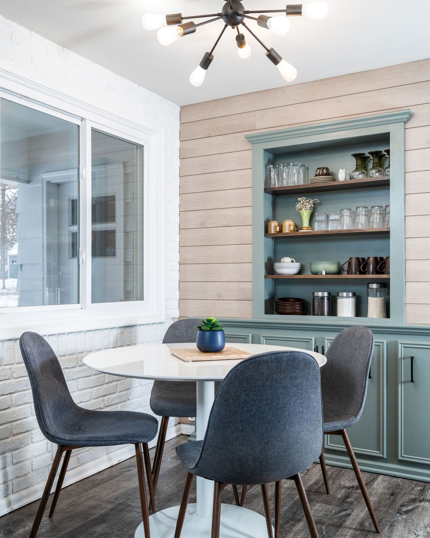 Modern dining area with round white table, gray upholstered chairs, and built-in sage green cabinet with open shelving.