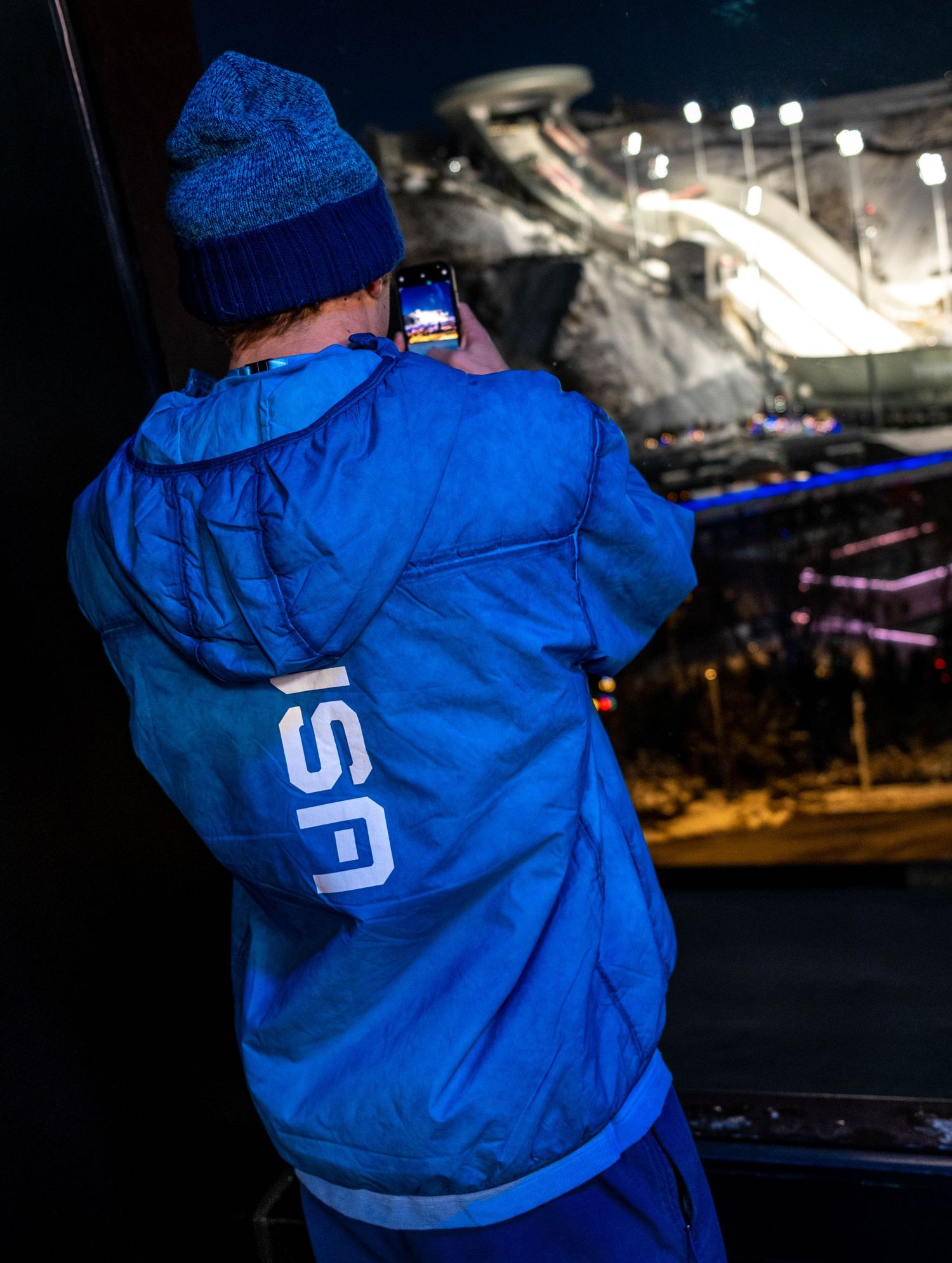 Person in USA team jacket taking photo of winter sports venue at night.