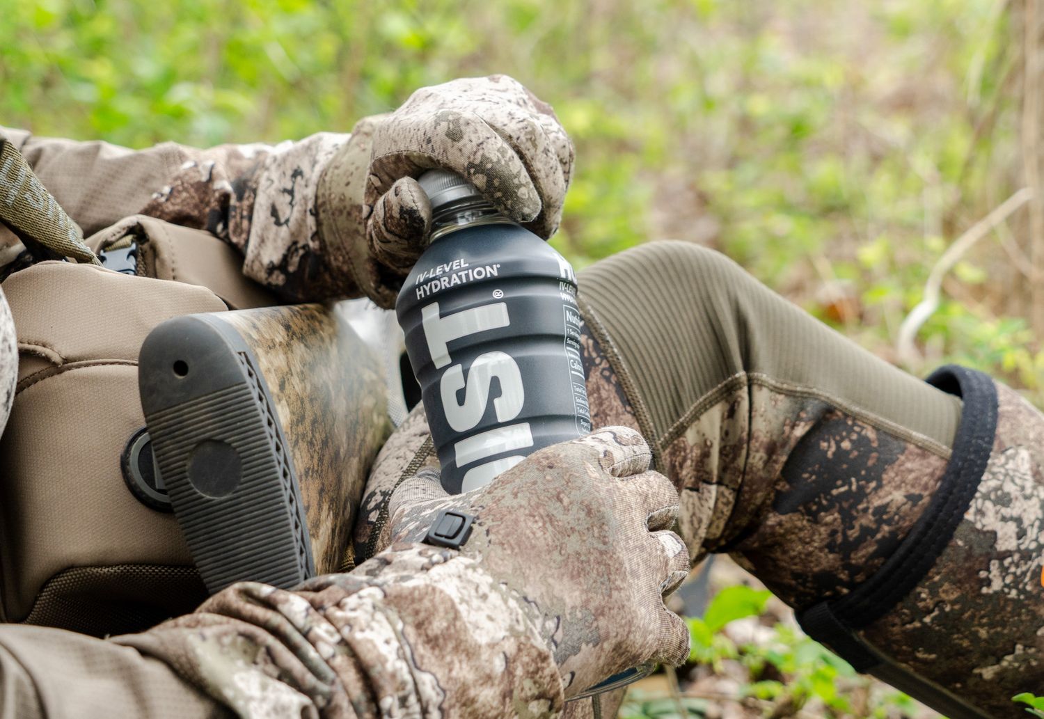 Camouflaged hunter holds black water bottle during outdoor activity.