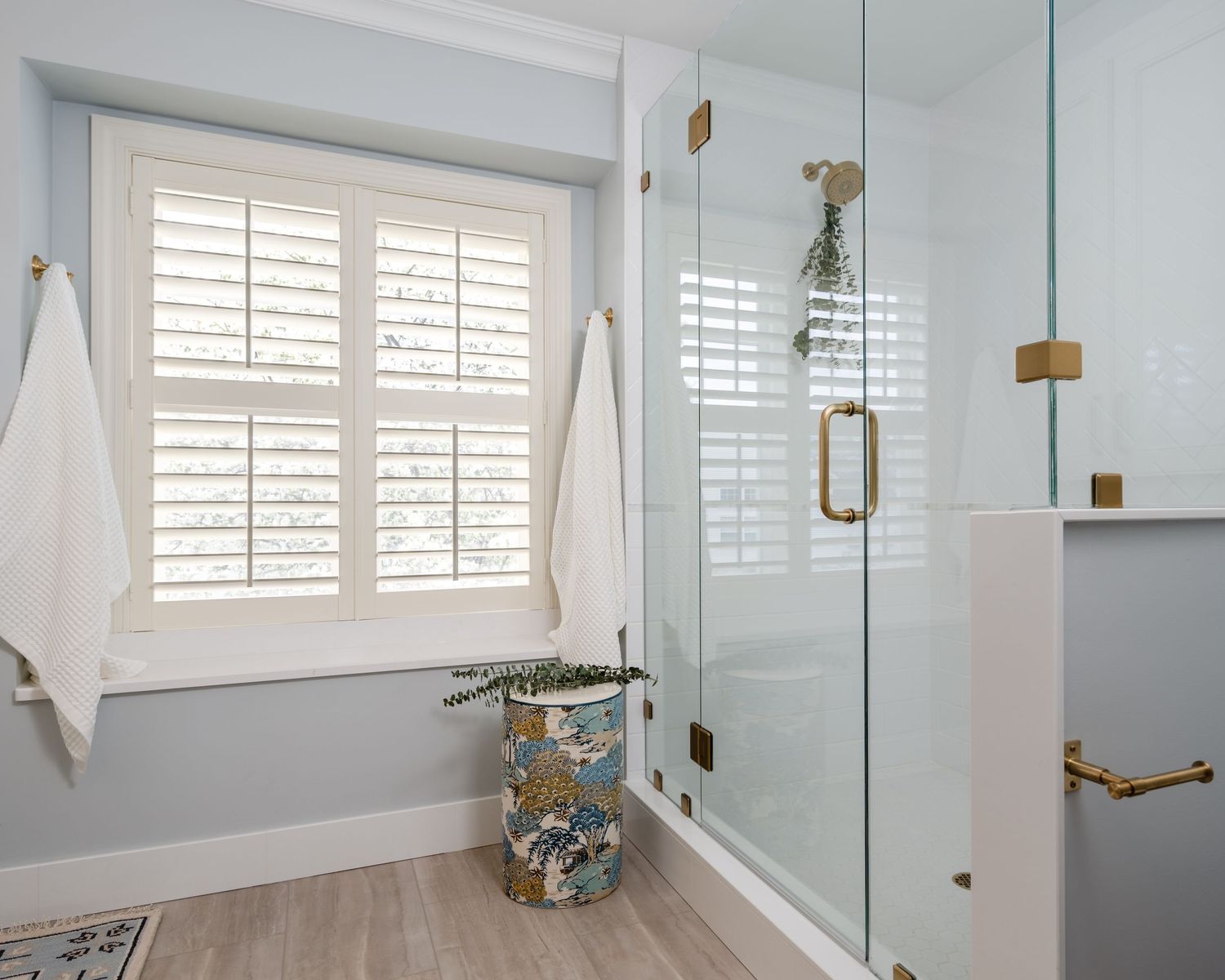 Modern bathroom with glass shower enclosure, brass fixtures, plantation shutters, and decorative floral waste bin on wood-look tile floor.