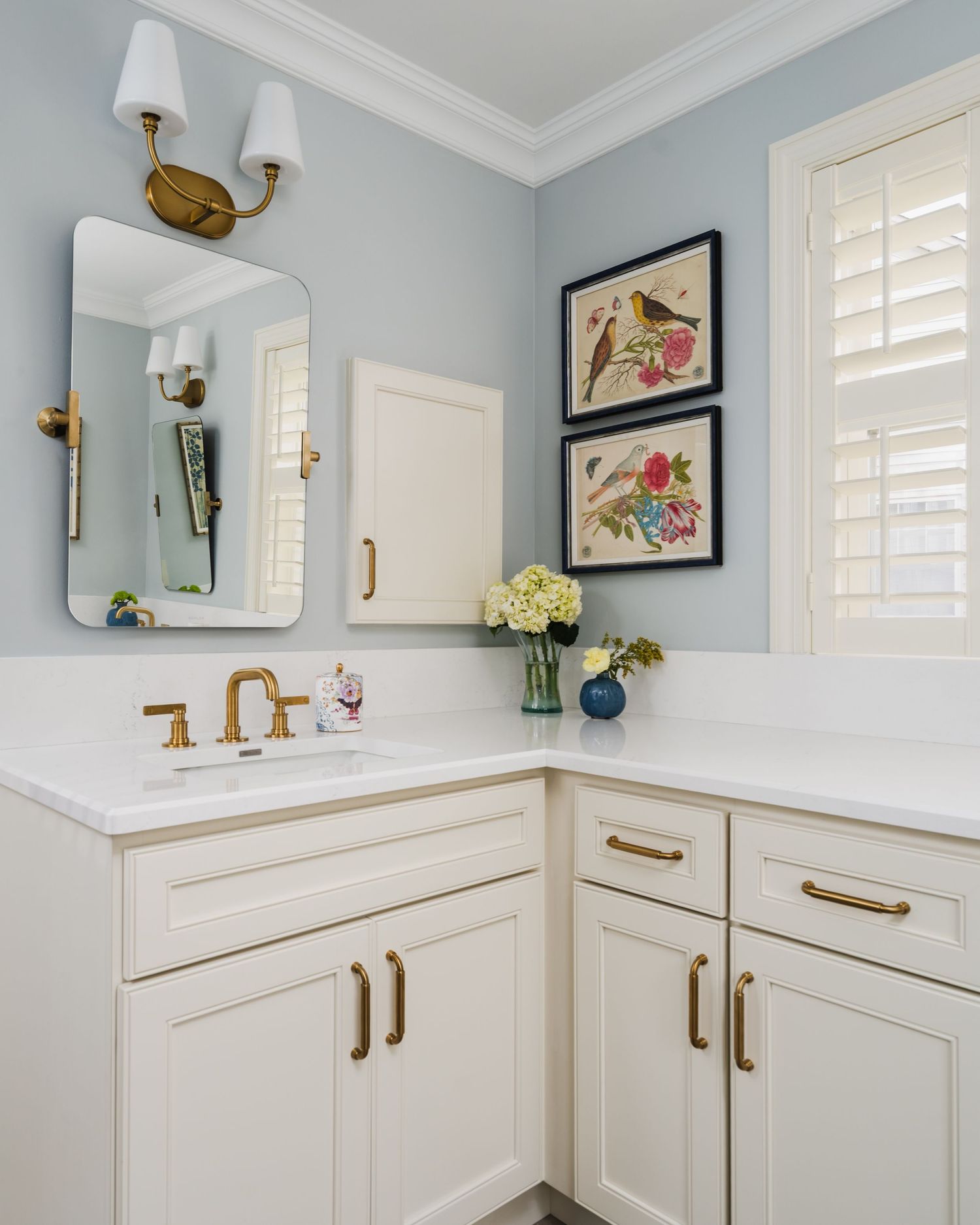 Elegant white bathroom vanity with gold hardware, blue-gray walls, arched mirror, and botanical artwork.