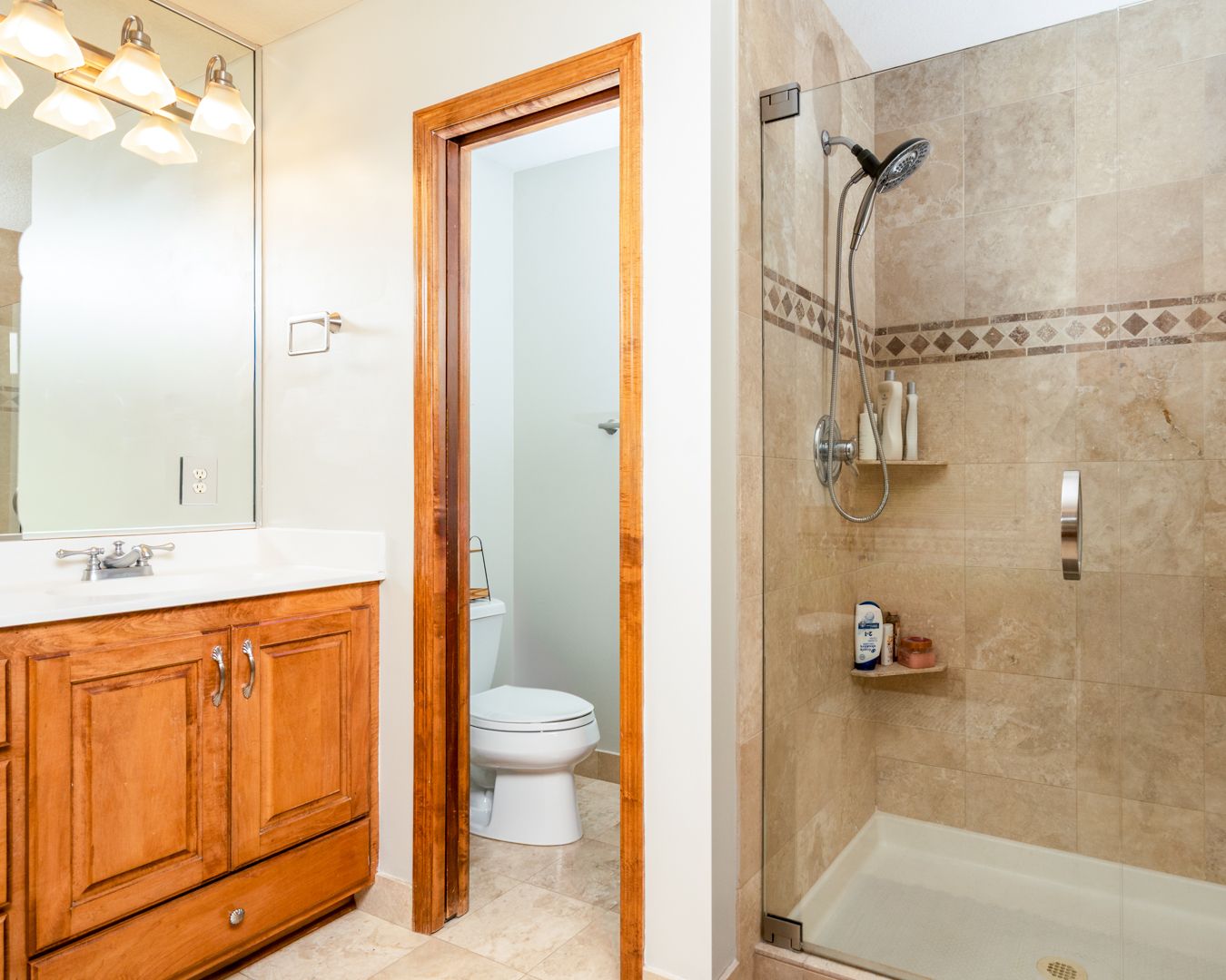 A modern bathroom interior with beige tiled walls, wooden cabinets, and glass shower enclosure shown in a panoramic view.