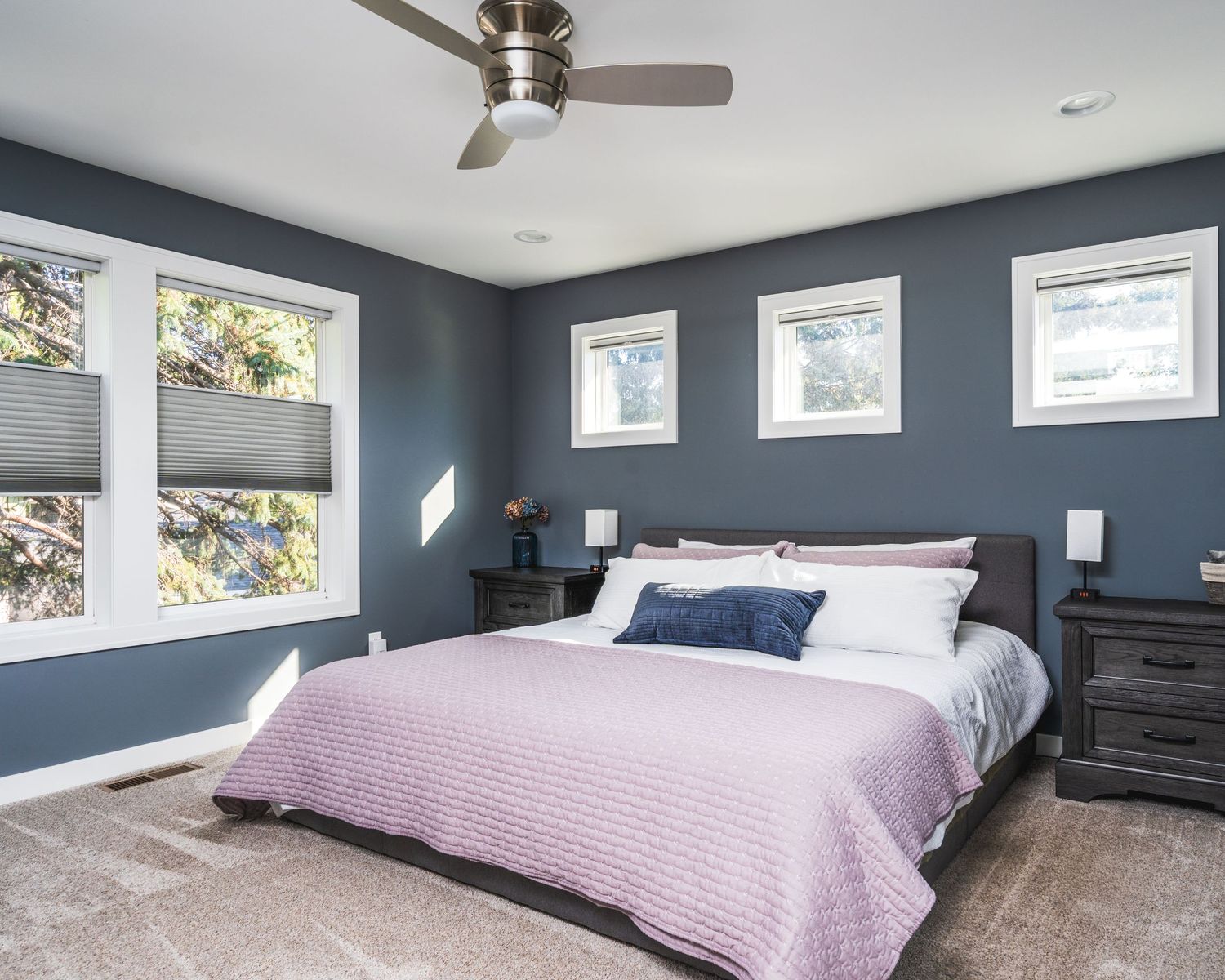 Modern bedroom with navy blue walls, large windows, hardwood floors, and a pink bedspread in natural lighting.