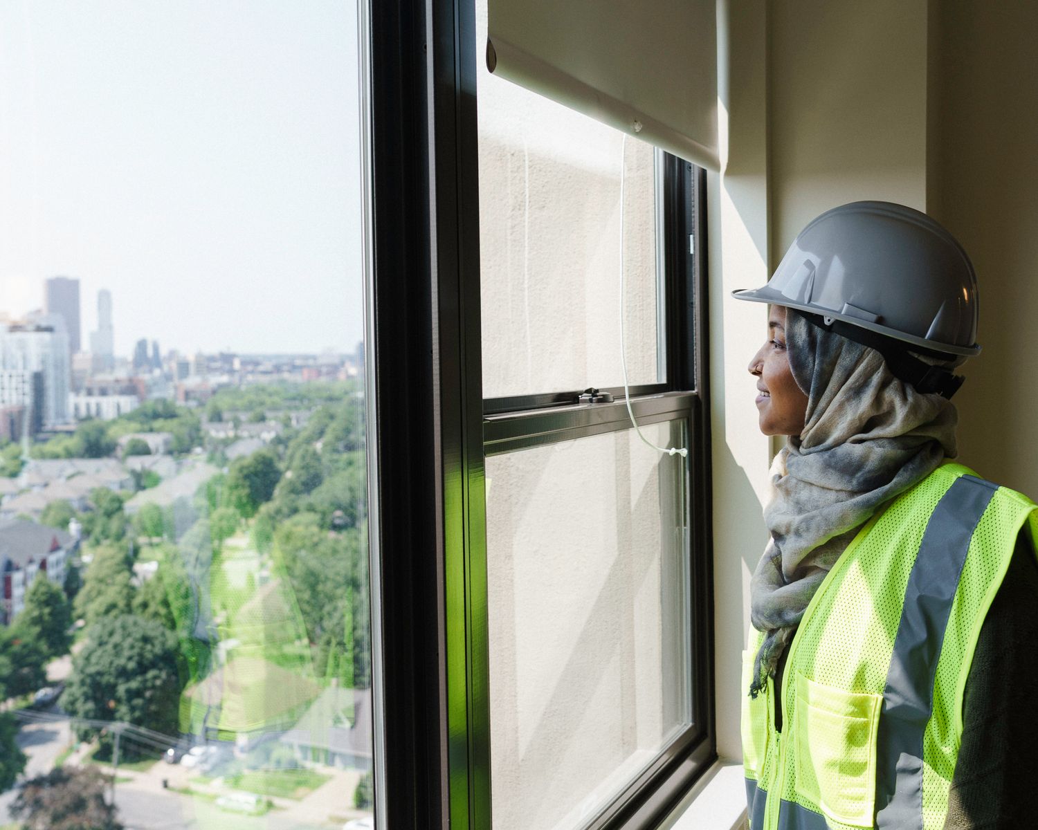 A female construction worker wearing a hijab, hard hat, and safety vest looks out a high-rise window at a city skyline.