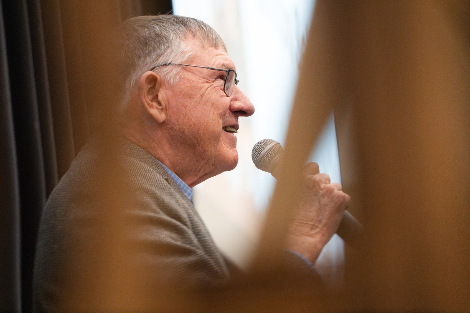 Person speaking into microphone at podium wearing glasses and gray sweater in profile view.