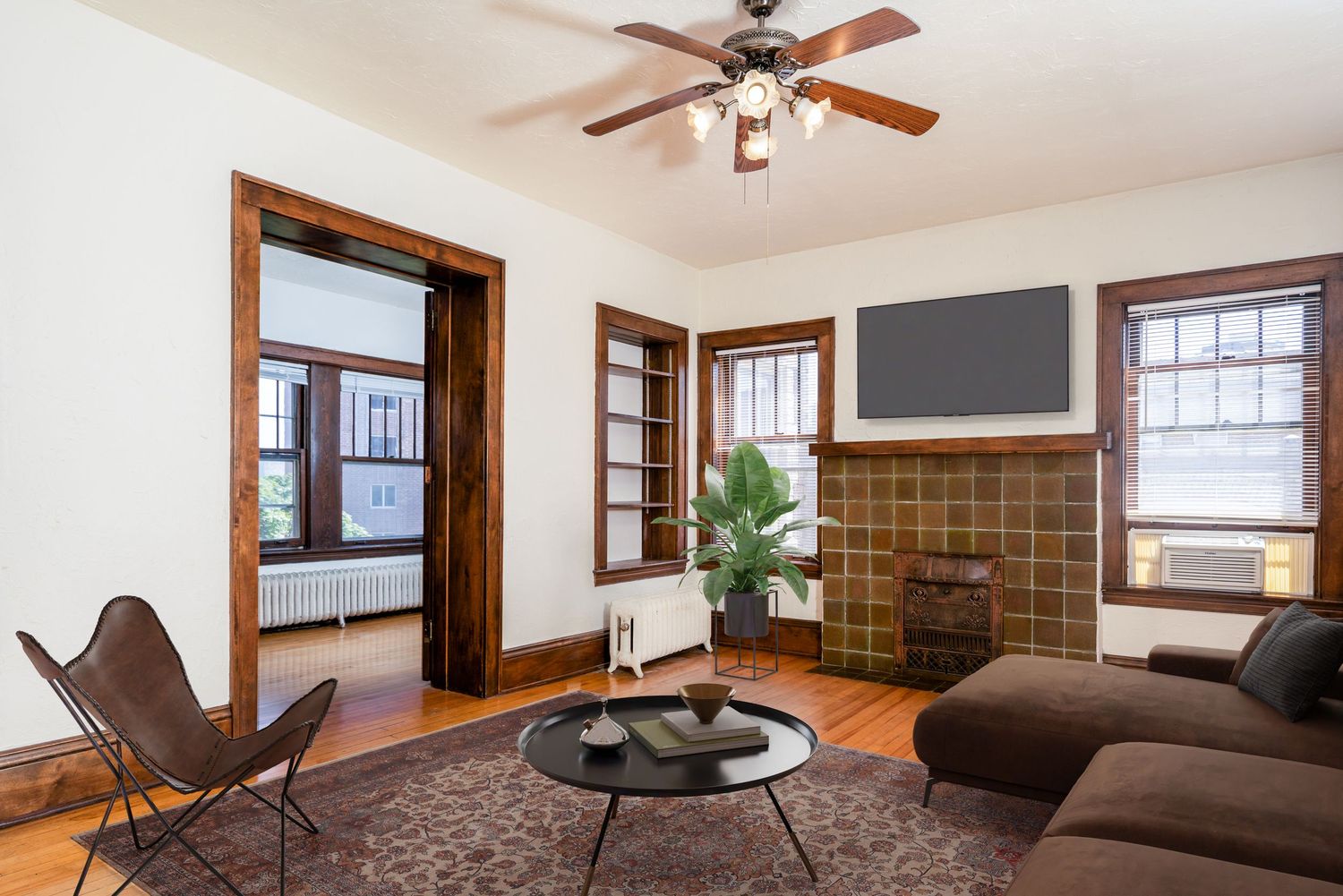 Modern living room with dark brown sofa, butterfly chair, ceiling fan, and brick fireplace against white walls.