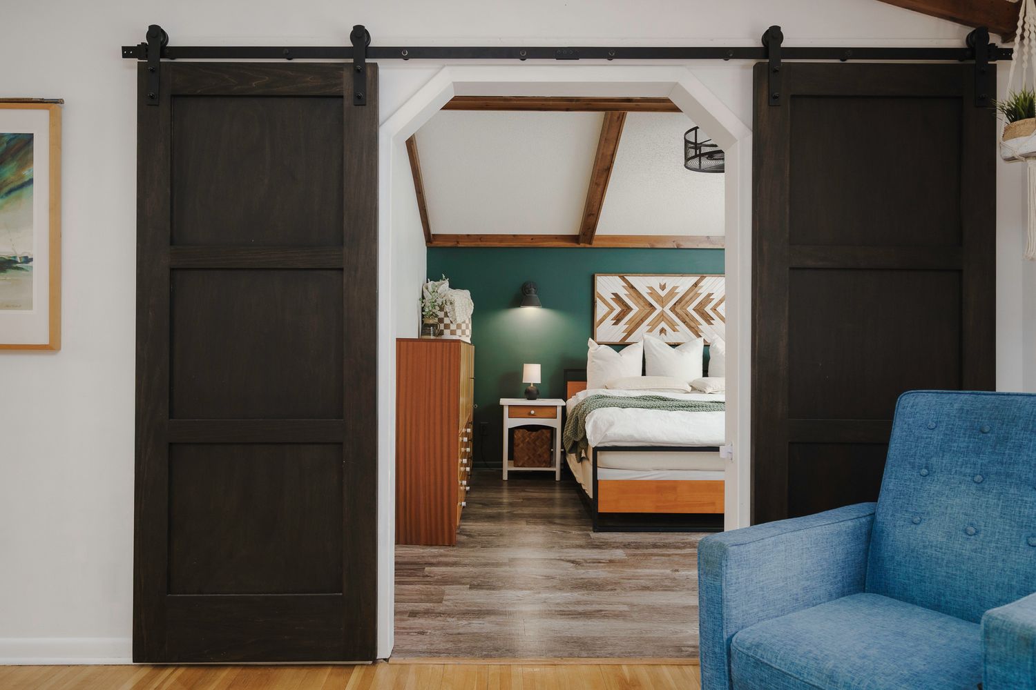Rustic hotel bedroom suite with dark sliding barn doors, geometric headboard, teal accent wall, and hardwood floors.