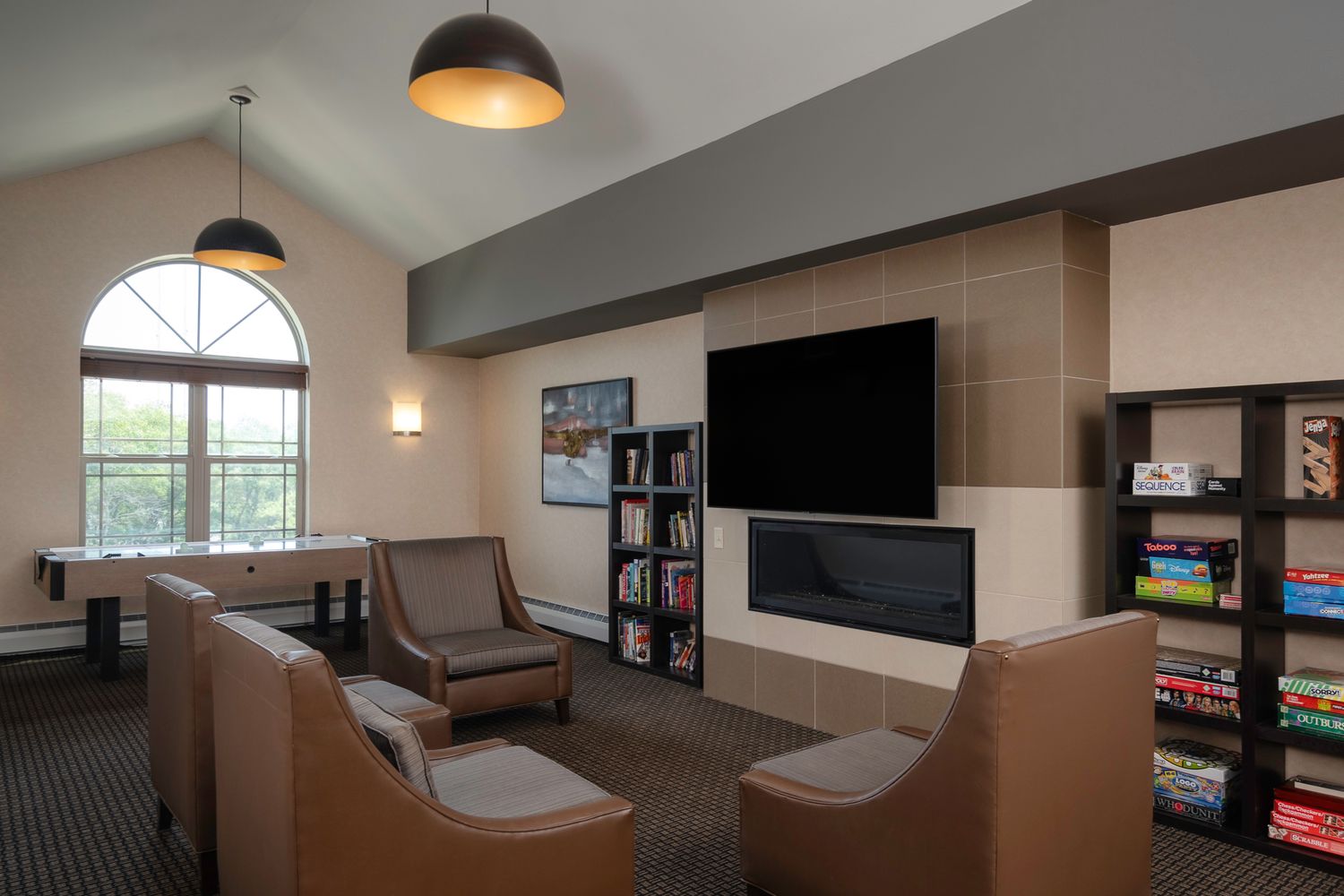 Cozy hotel lounge with leather chairs, wall-mounted TV, fireplace, bookshelves, and arched window with natural light.