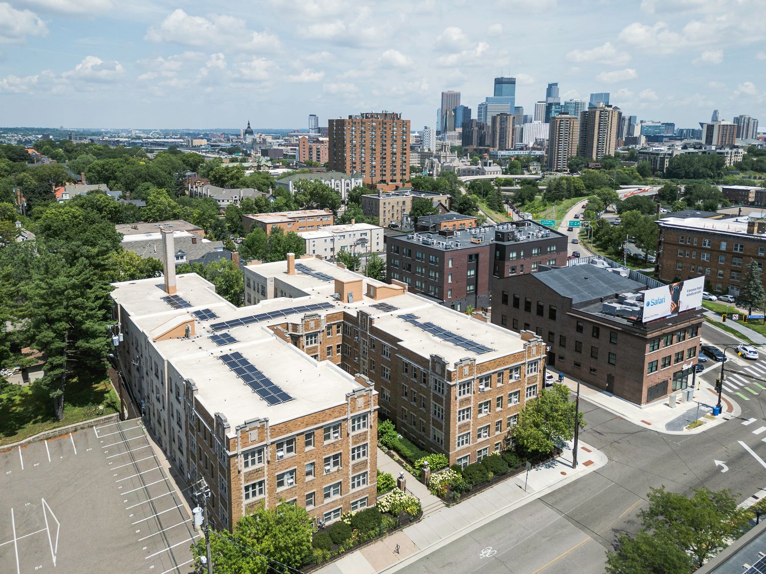 Aerial view of brick apartment complex with solar panels near Minneapolis skyline surrounded by lush green trees.