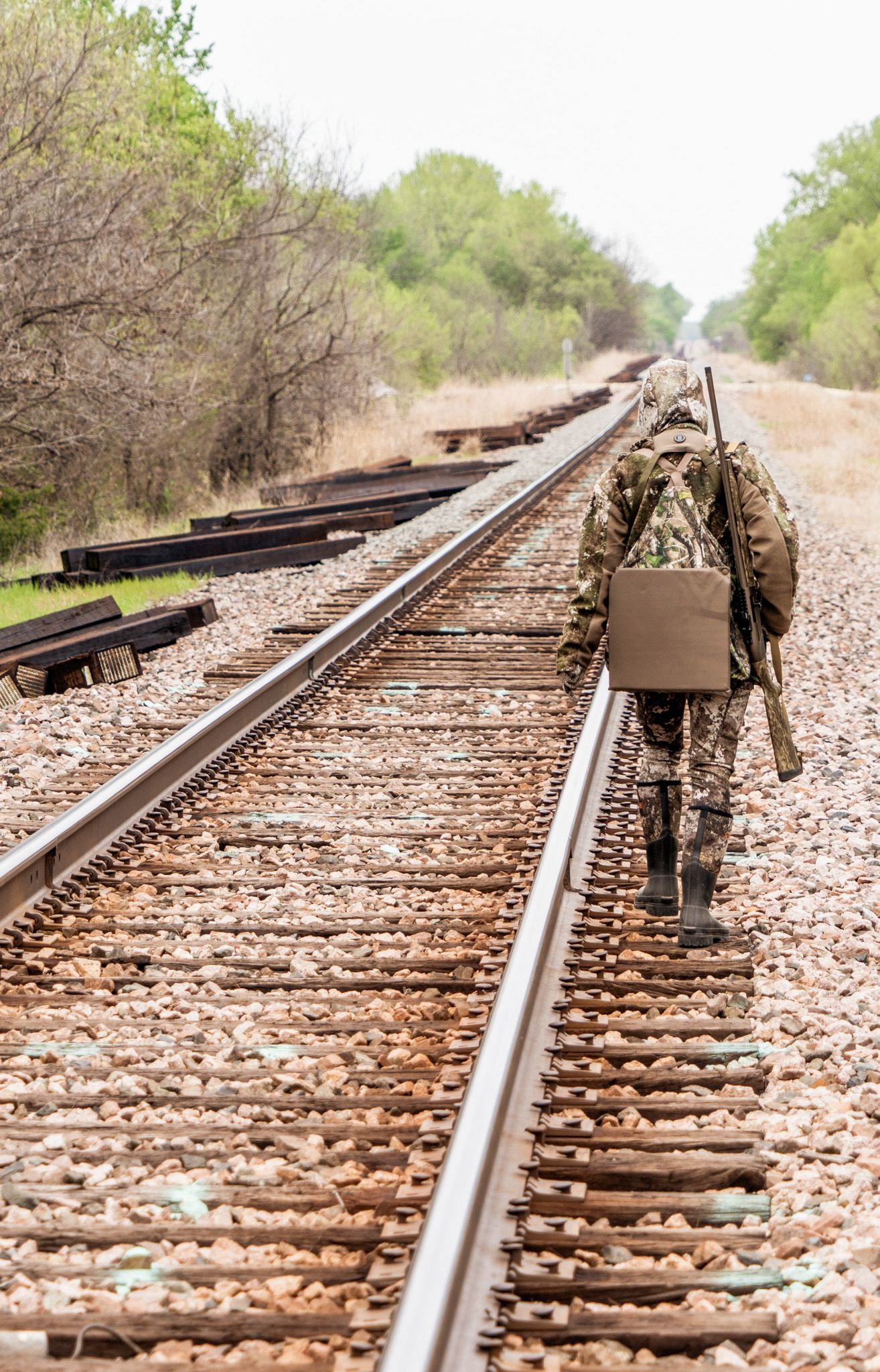 Hunter in camouflage gear walks along railroad tracks through rural landscape.