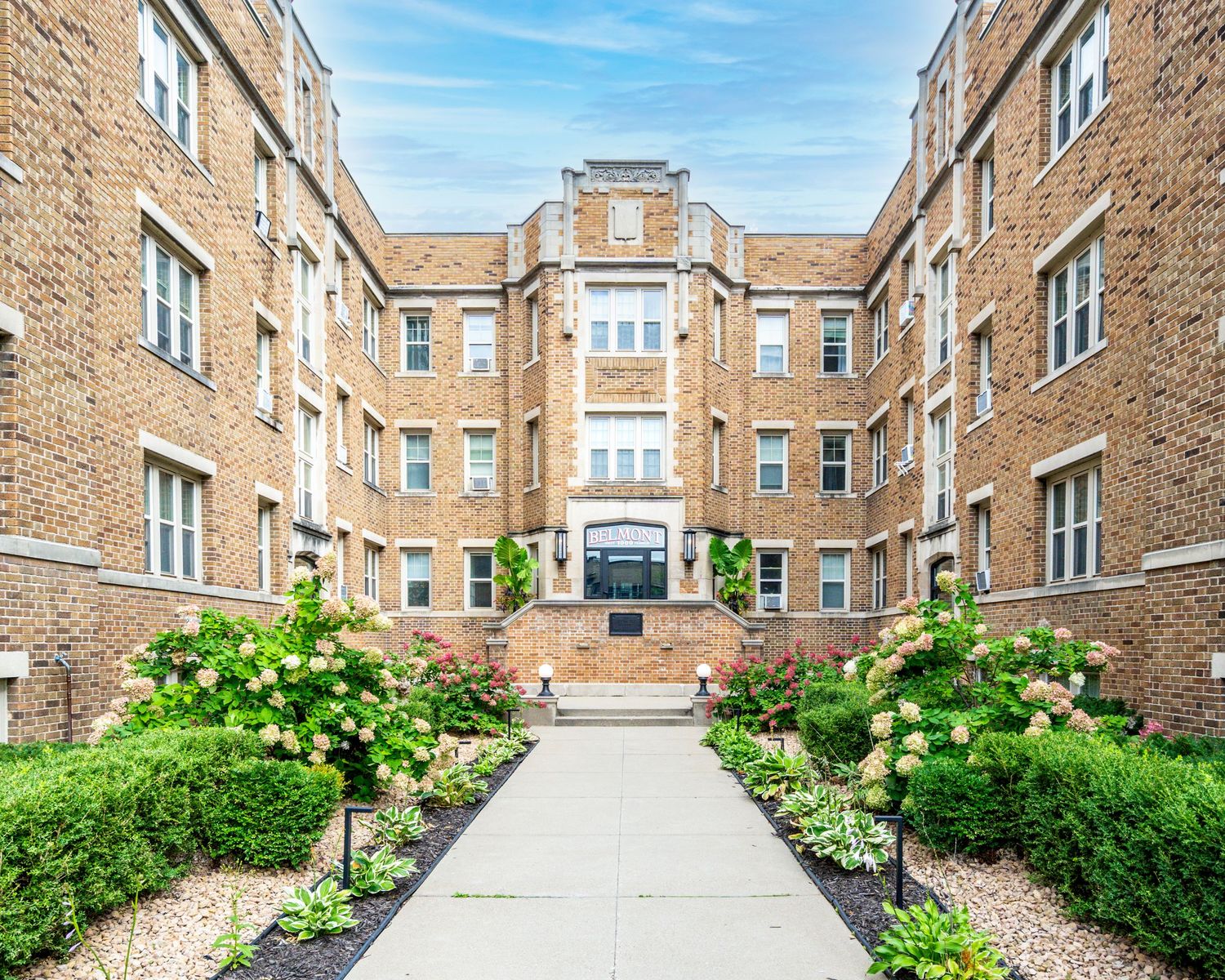 Charming brick apartment building with manicured courtyard, lush hydrangeas, and a welcoming entrance pathway on a sunny day.