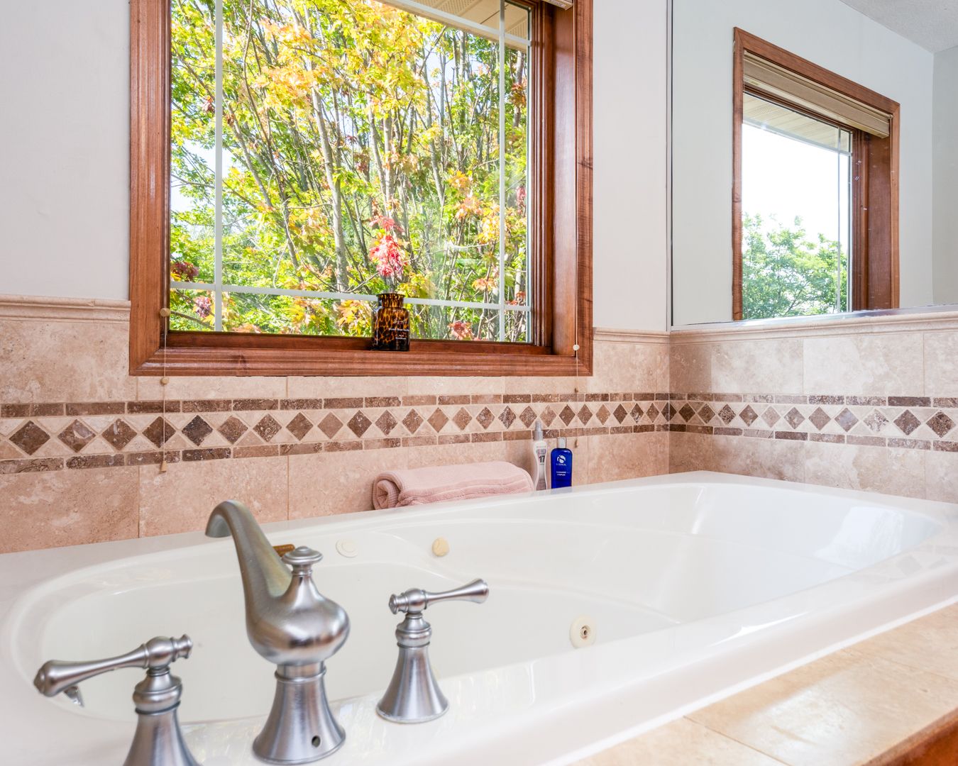 Luxury bathroom features white soaking tub with chrome fixtures beneath window overlooking lush autumn trees outside.