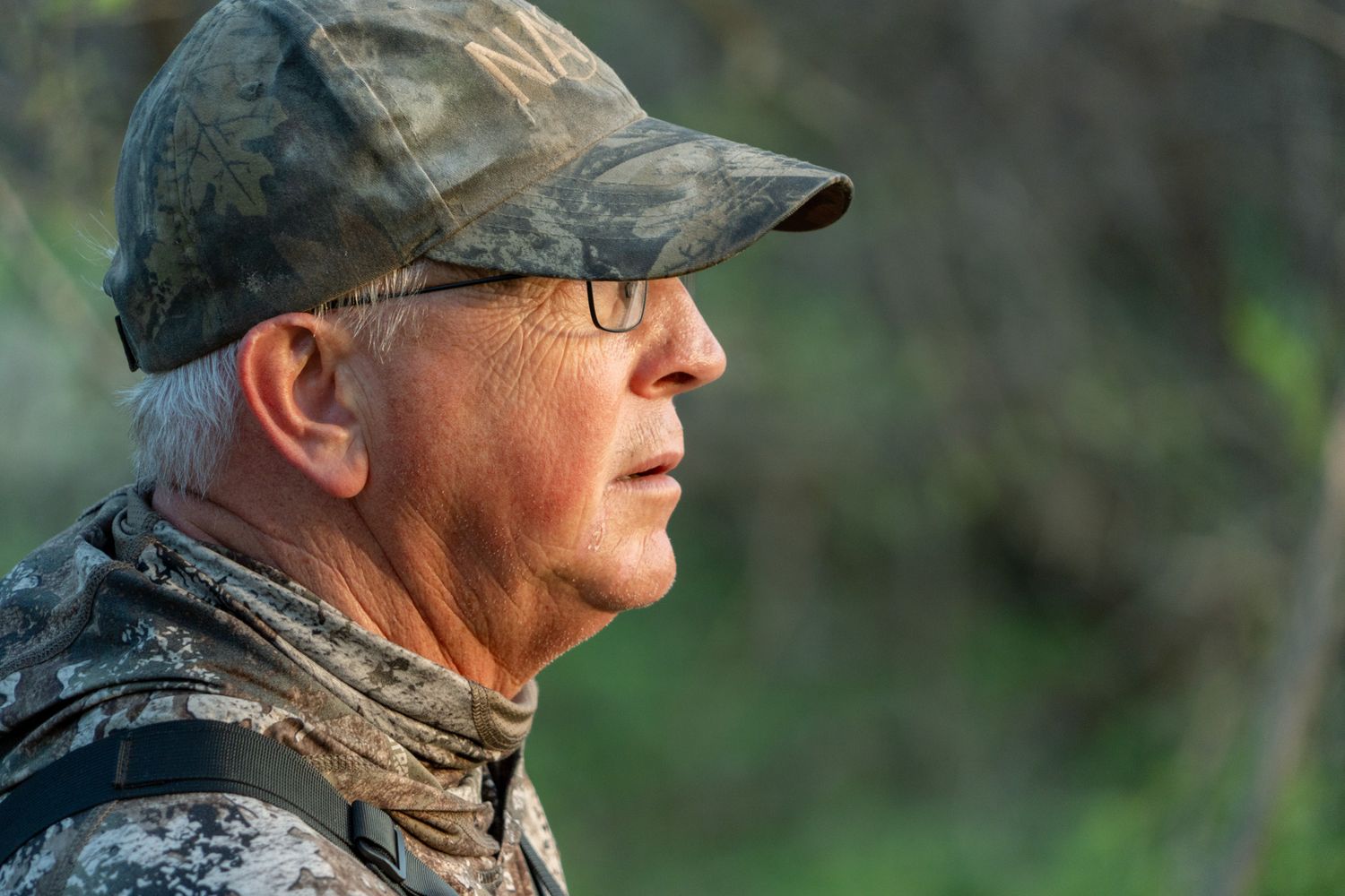 Person in camo hunting gear wears glasses and cap in profile view.