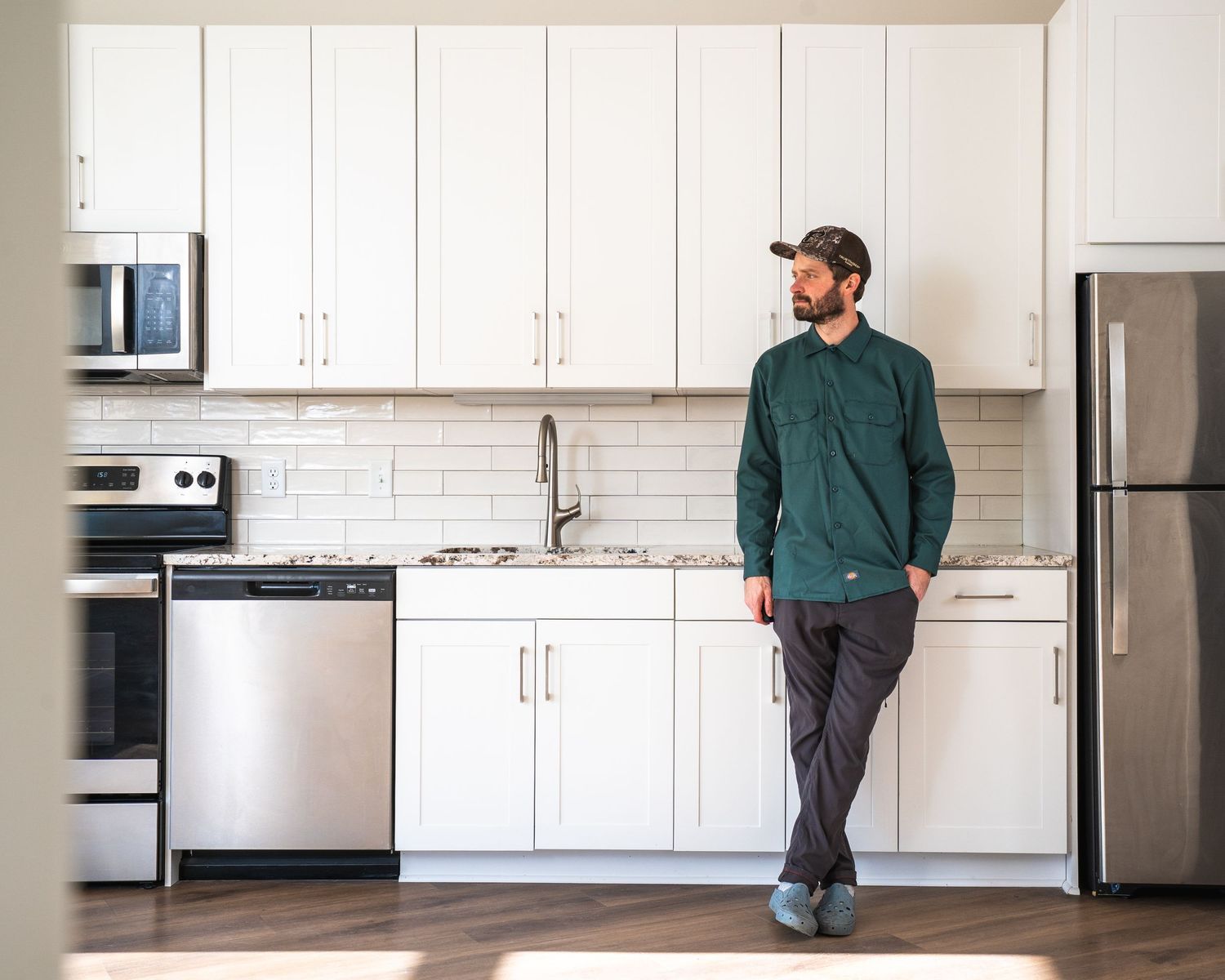 Man in green shirt and cap standing in modern white kitchen with stainless steel appliances and subway tile backsplash.