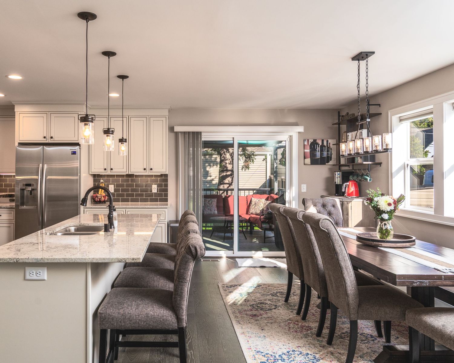 Modern open concept kitchen and dining area with marble countertops, pendant lighting, and gray upholstered seating.