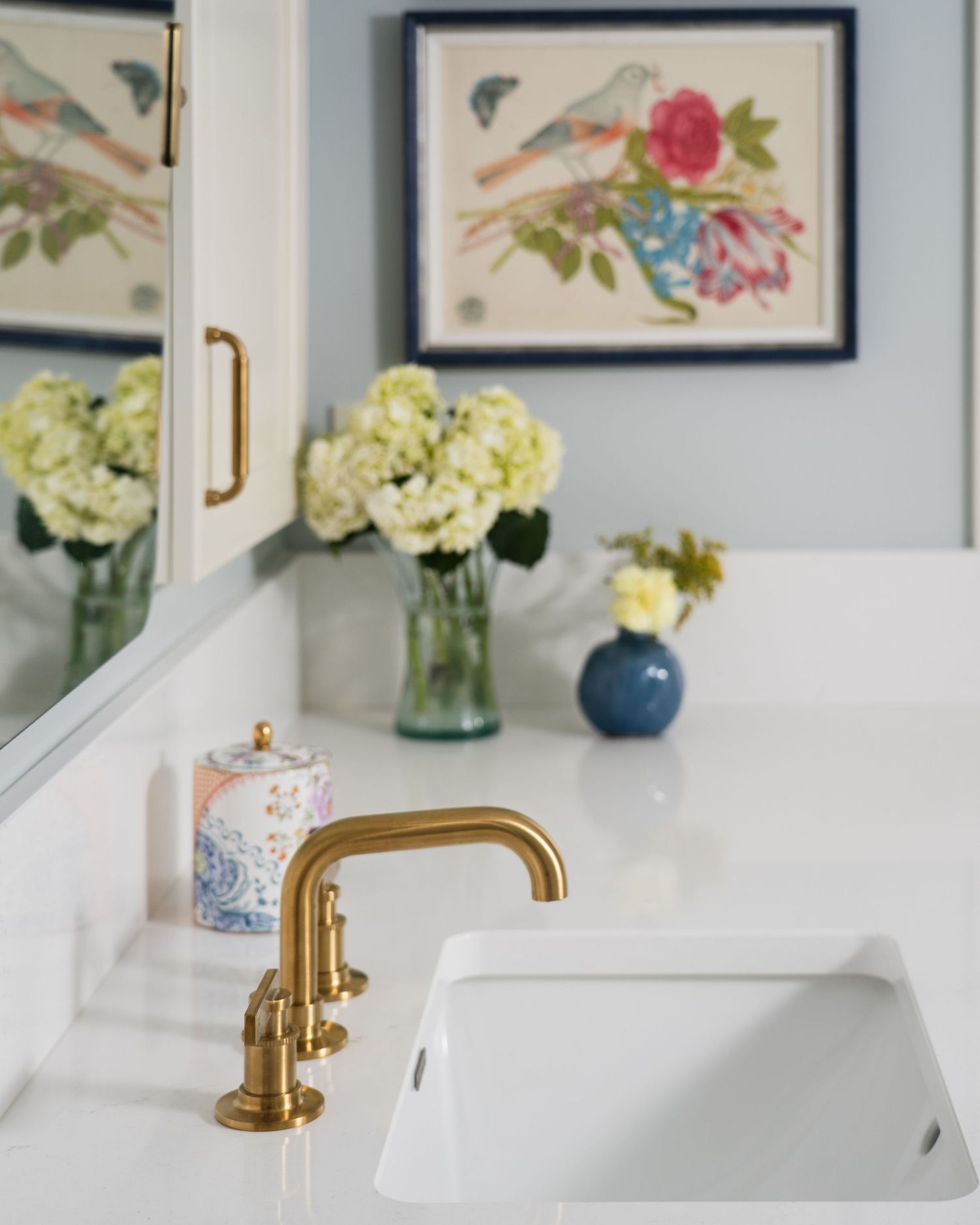 Elegant bathroom vanity with gold faucet, white sink, hydrangea flowers, and colorful bird art on blue walls.