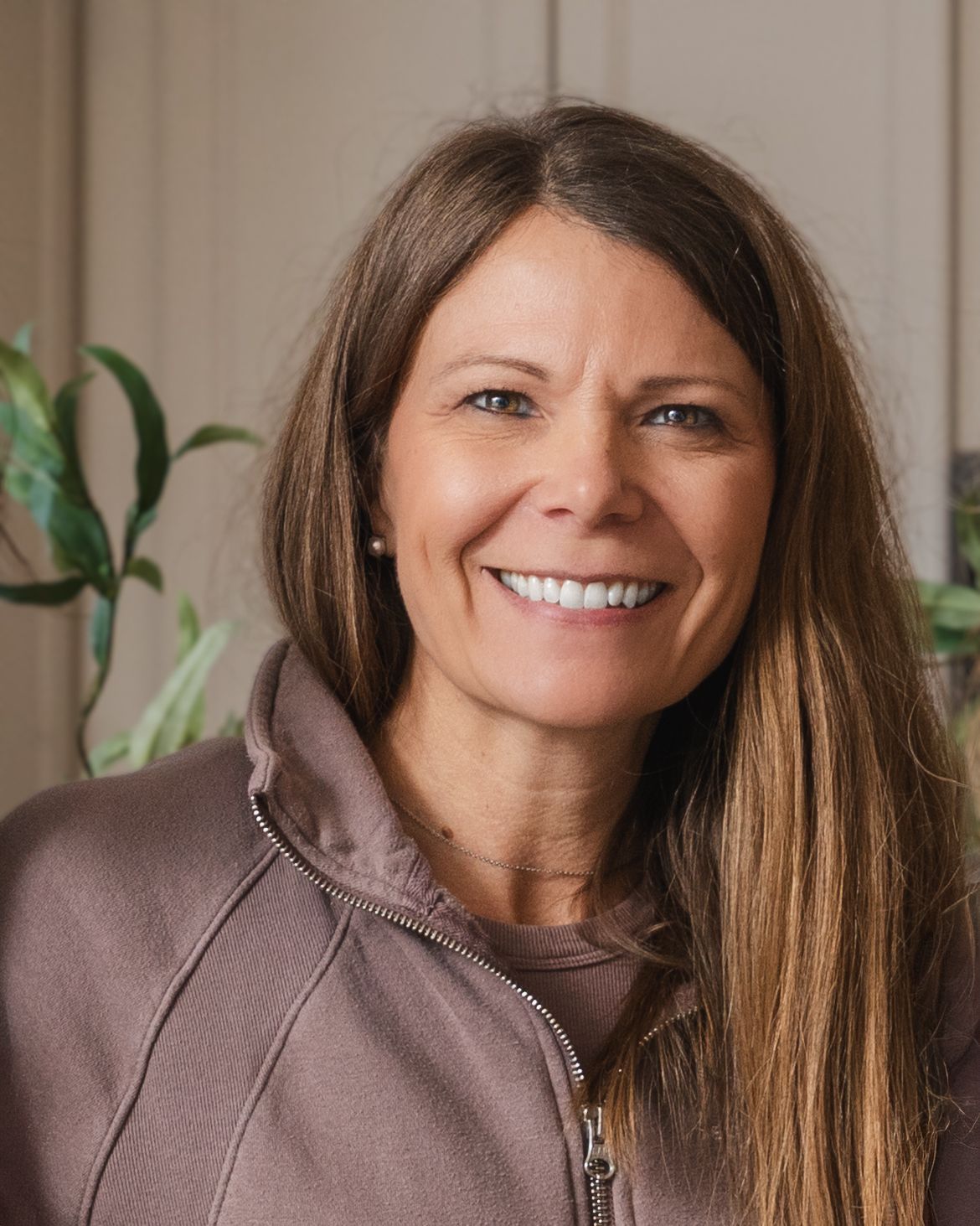 Professional headshot of someone wearing a brown zip-up jacket, with a warm smile and long brown hair against a light background.