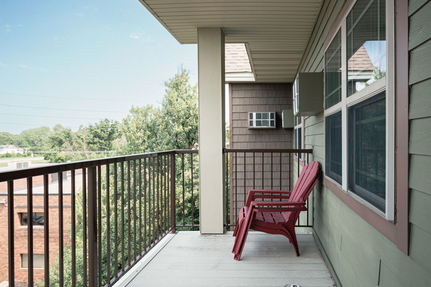 A red Adirondack chair on a second-floor apartment balcony with iron railings overlooking green trees.