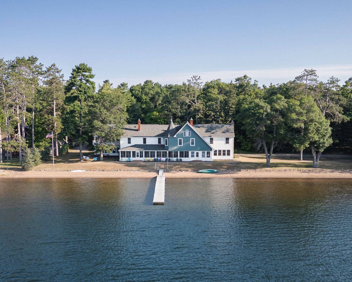 Aerial view of a large waterfront estate with a private dock, sandy beach, and lush trees under a clear blue sky.