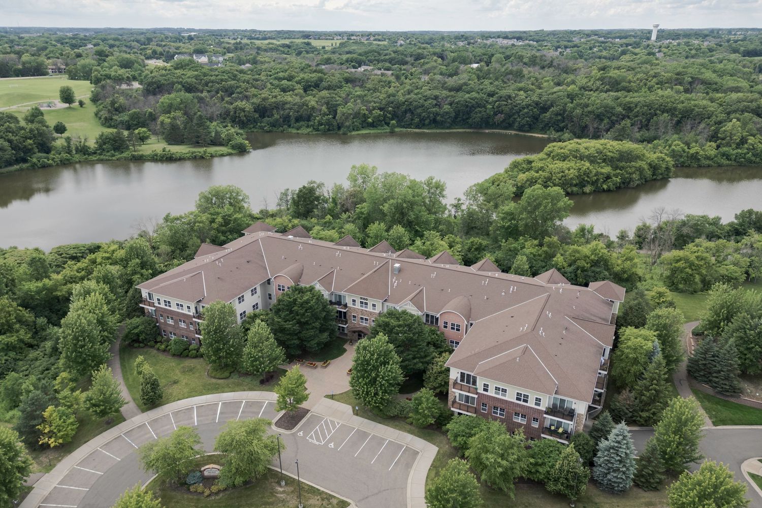 Aerial view of a brick apartment complex surrounded by lush trees and a serene lake in a suburban setting.