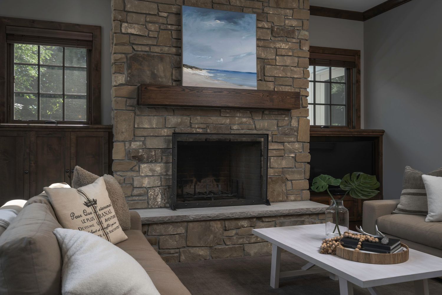 Cozy living room with stone fireplace, wood mantel, neutral sofa, white coffee table, and coastal painting above hearth.