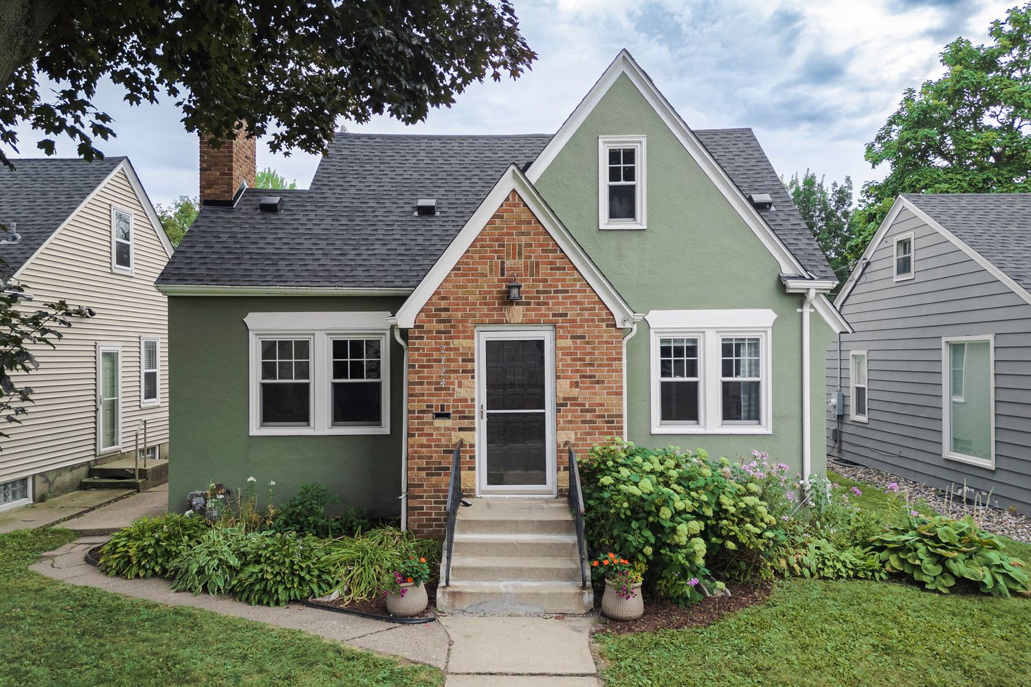 Green Cape Cod home with brick entryway, white trim, lush landscaping, and charming curb appeal on a sunny day.