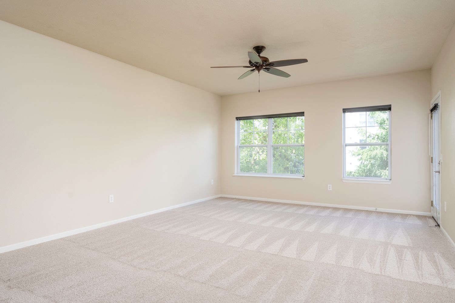 Bright empty living room with ceiling fan, neutral carpet, and two large windows with natural light.