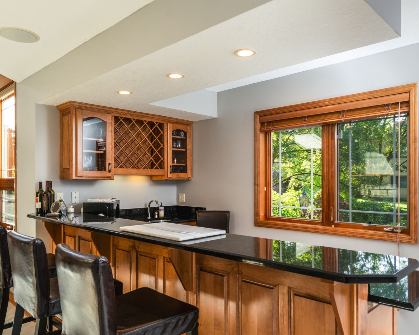 Modern kitchen interior featuring granite countertops, wooden cabinets and recessed lighting with forest views through windows.