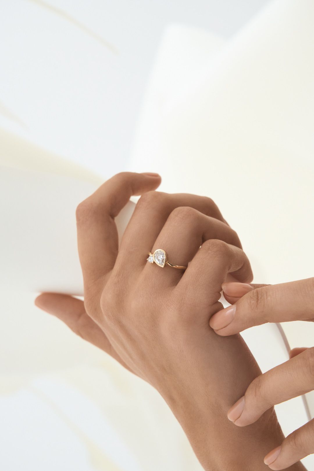 Close-up of hands wearing a delicate solitaire diamond engagement ring on a light background.