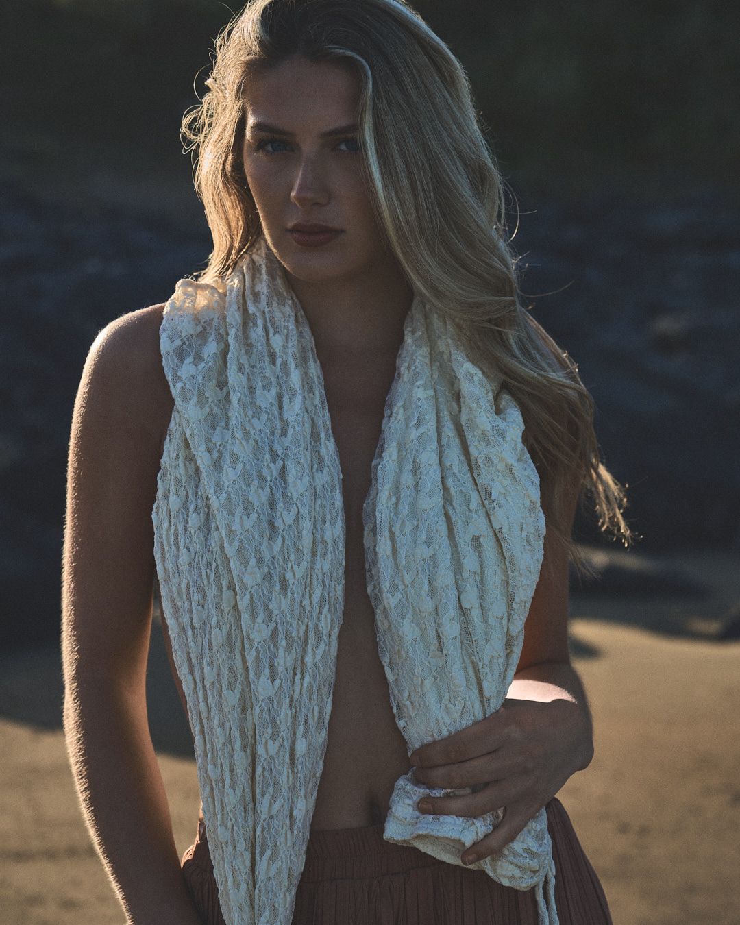 Figure draped in textured white scarf poses on beach at golden hour.